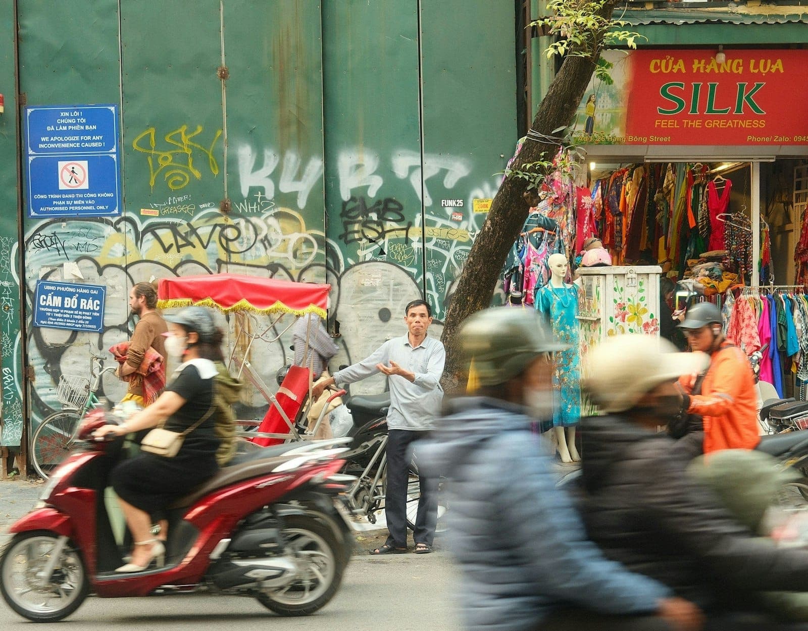 Scène de rue animée à Hanoï avec des personnes à moto et un cyclo devant des commerces locaux aux devantures colorées.