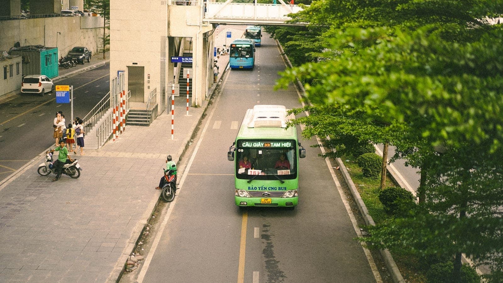 Bus public vert roulant dans une rue à Hanoi avec un autre bus au loin, entouré d'arbres et de bâtiments.