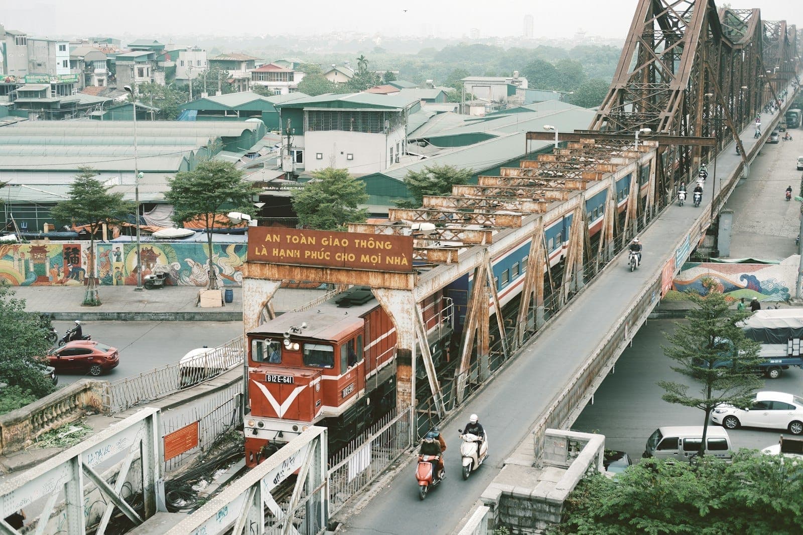 Un train de voyageurs traversant l'emblématique pont Long Bien de Hanoi avec des bâtiments de la ville et des motos en contrebas, illustrant les itinéraires de départ pour des excursions d'une journée.
