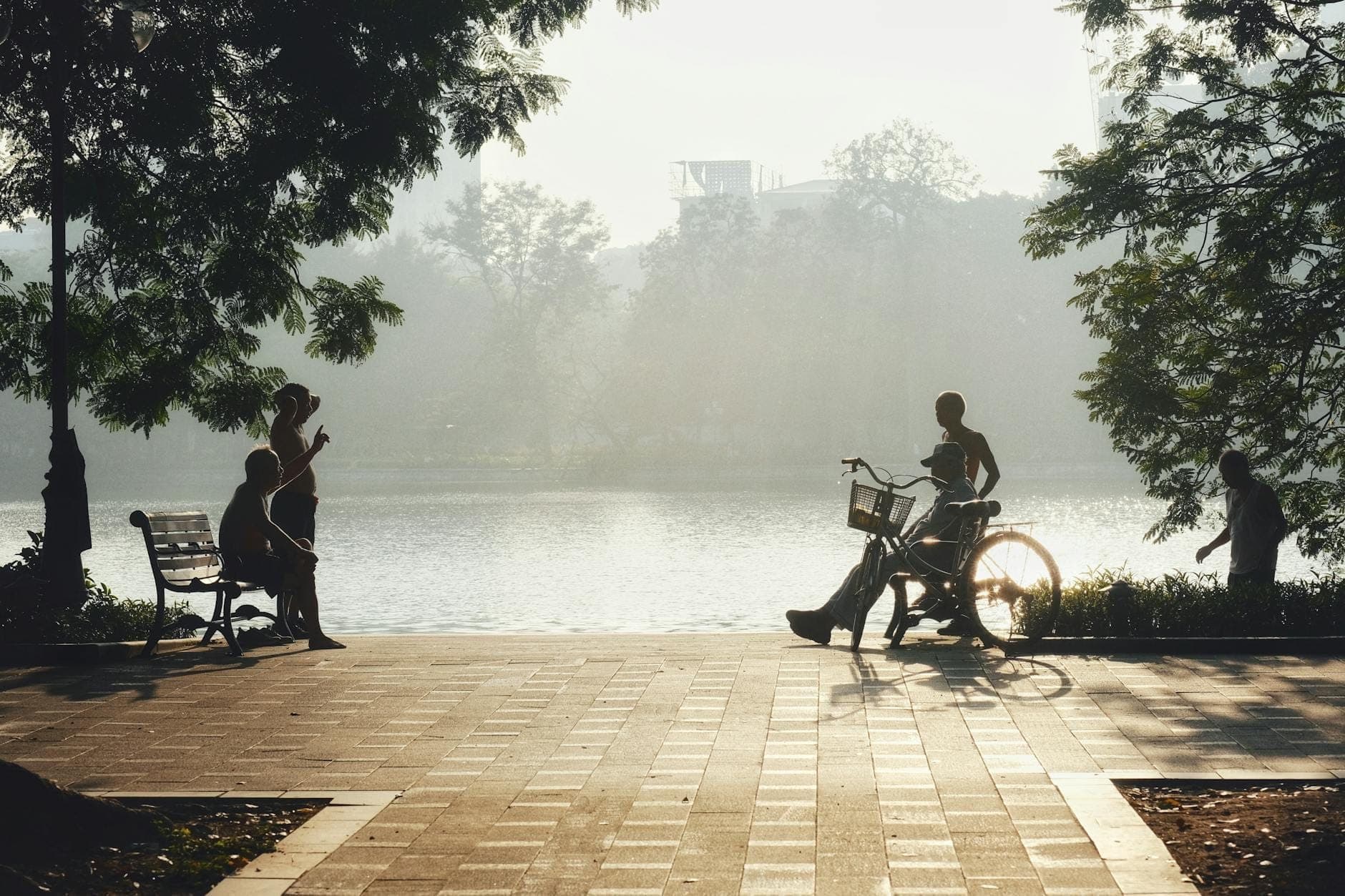 Des personnes marchant, assises et faisant du vélo le long d'un chemin pavé en bord de lac ombragé par des arbres, avec une lumière brumeuse sur l'eau à Hanoi.