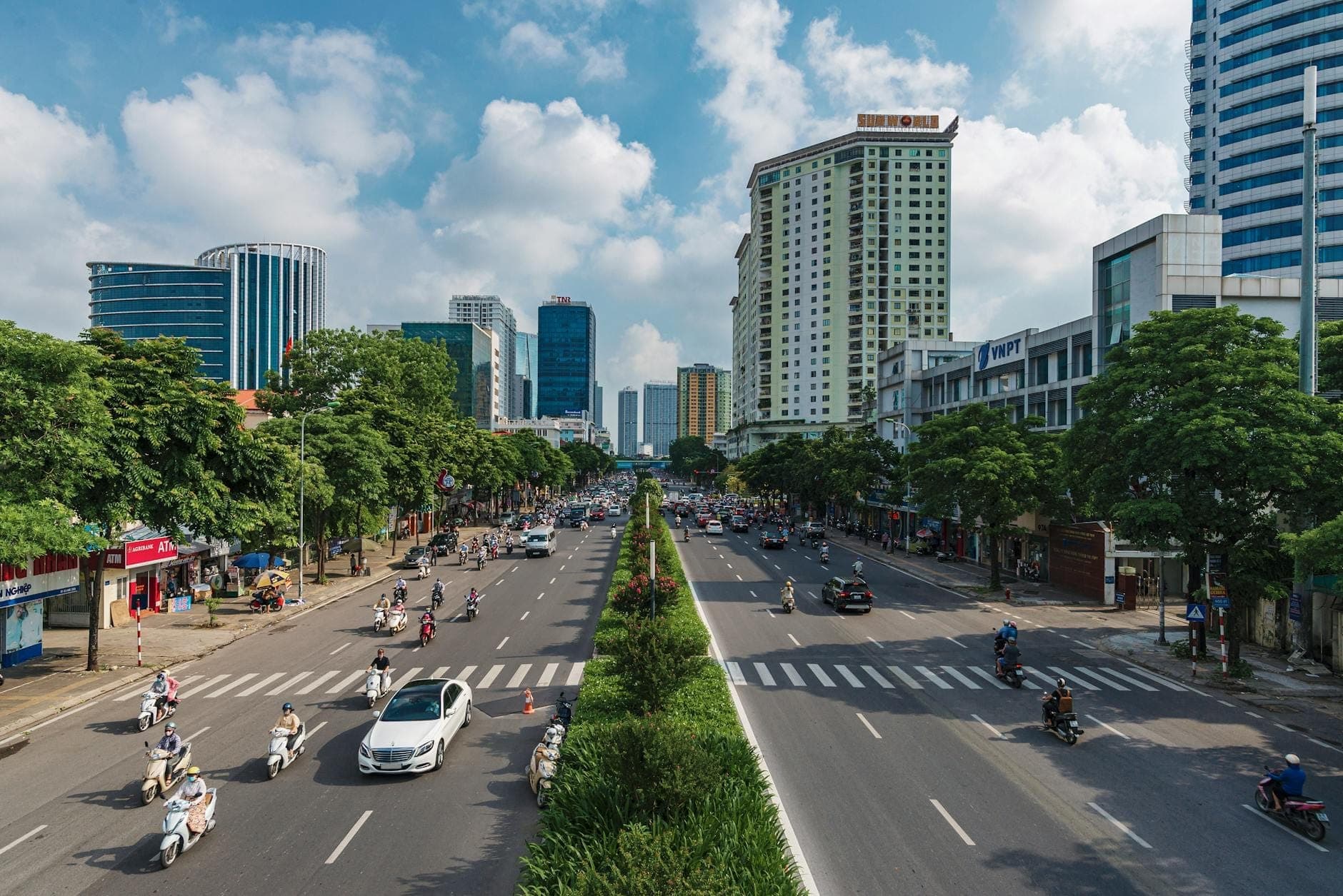 Large boulevard à Hanoi avec des voitures et des motos circulant entre les quartiers, entouré de bâtiments modernes et d'arbres sous un ciel lumineux.
