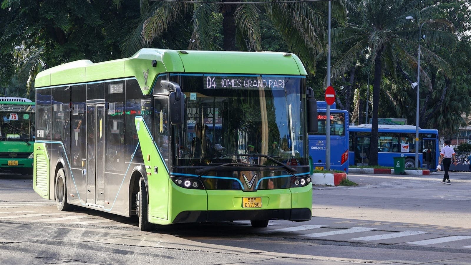 A green public bus in Ho Chi Minh City at a street intersection with other buses and palm trees in the background.