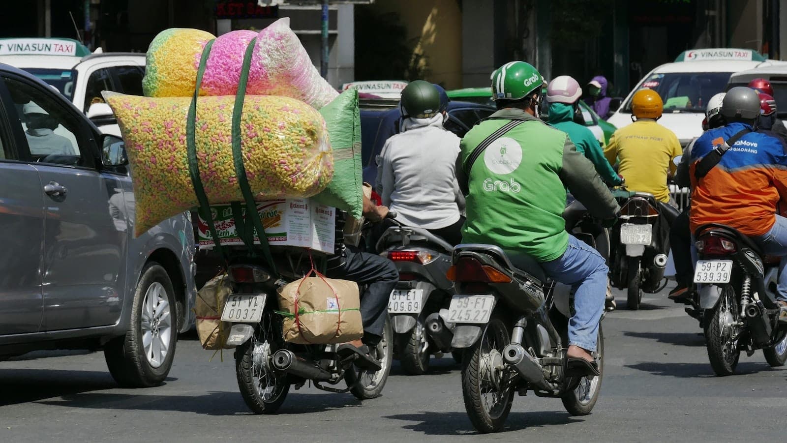 Multiple motorbike drivers, including a Grab rider in green jacket and helmet, and taxis in traffic on a busy Ho Chi Minh City street.