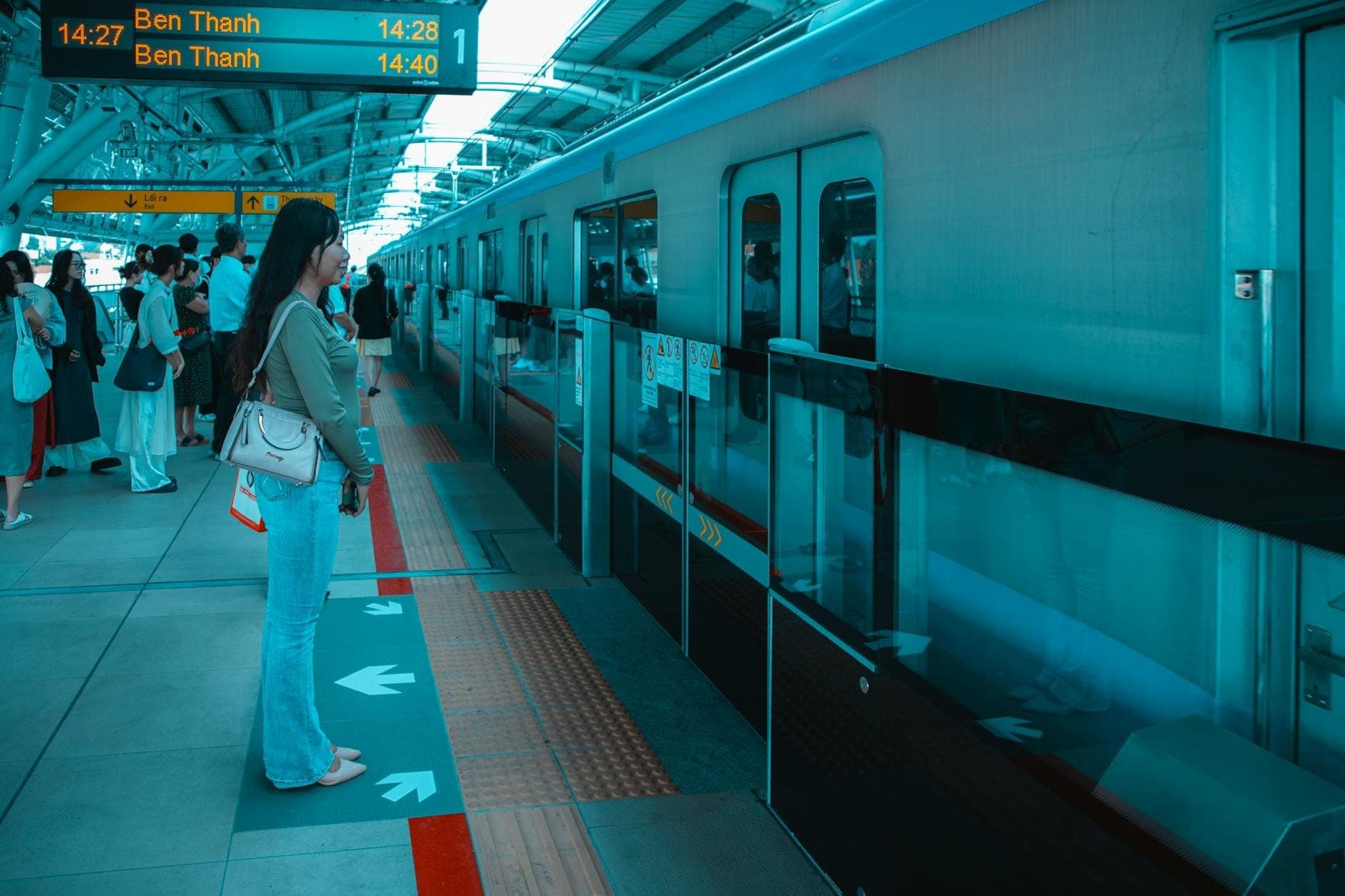 Passengers standing on a modern metro platform in Ho Chi Minh City, waiting for a train to Ben Thanh station, with clear signage overhead.