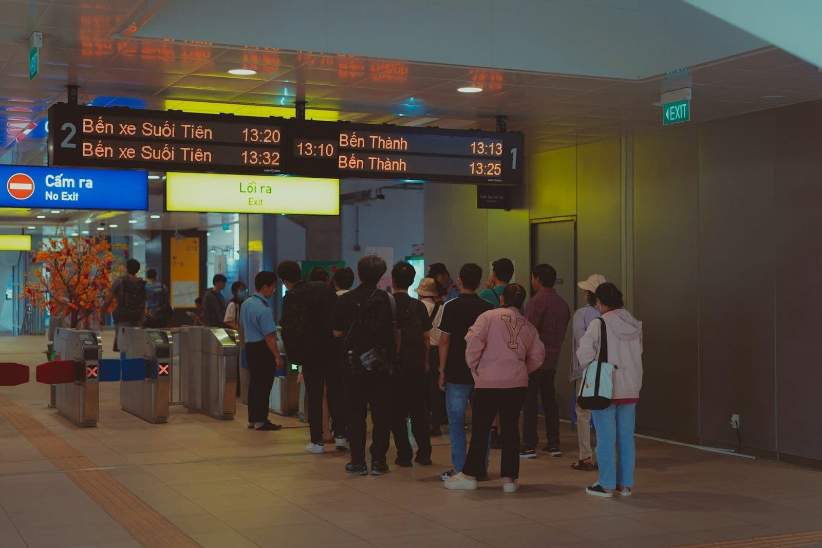 Travelers queuing at a brightly lit metro or rail station in Ho Chi Minh City, with visible signs to Suoi Tien and Ben Thanh terminals.