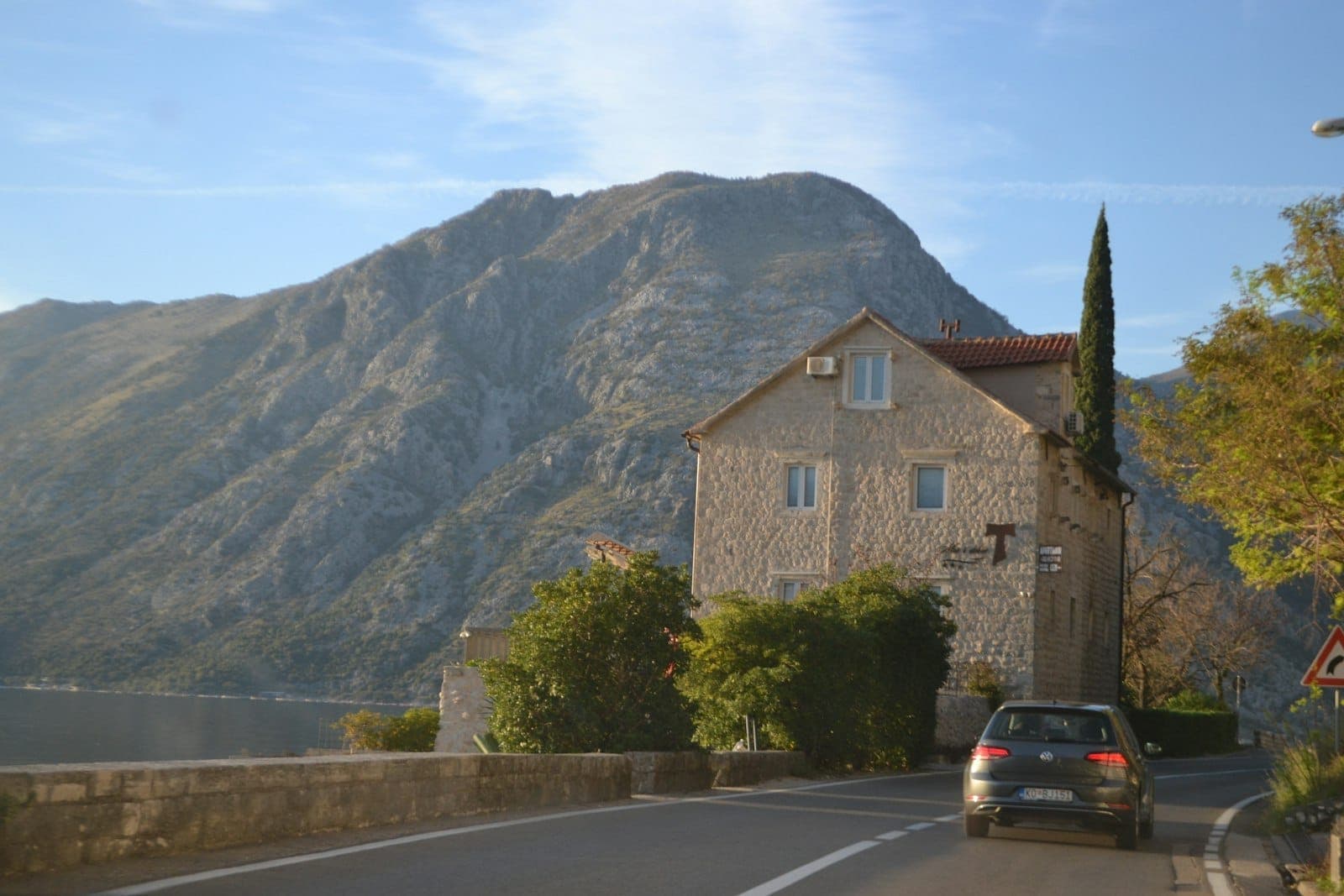 Une voiture roule le long d'une route côtière à Kotor, longeant des maisons en pierre avec une montagne en arrière-plan et la baie visible à proximité.