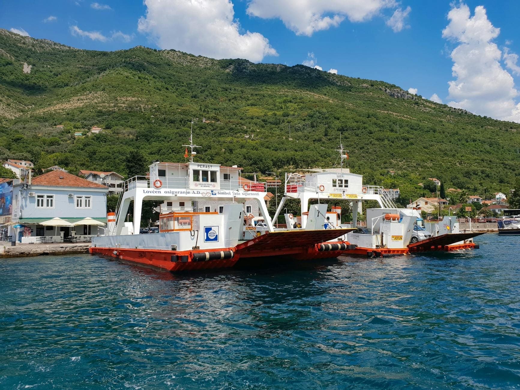 Ferries pour voitures amarrés dans un port de la Baie de Kotor avec des collines verdoyantes et des bâtiments en arrière-plan.