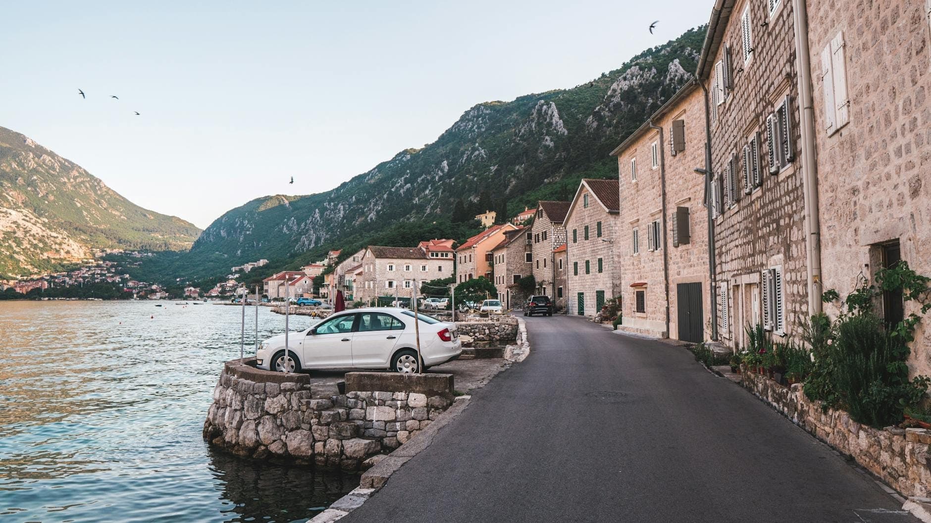 Des bâtiments en pierre longent une route calme au bord de l'eau dans la Baie de Kotor, avec une voiture garée près de l'eau et des montagnes verdoyantes en arrière-plan.