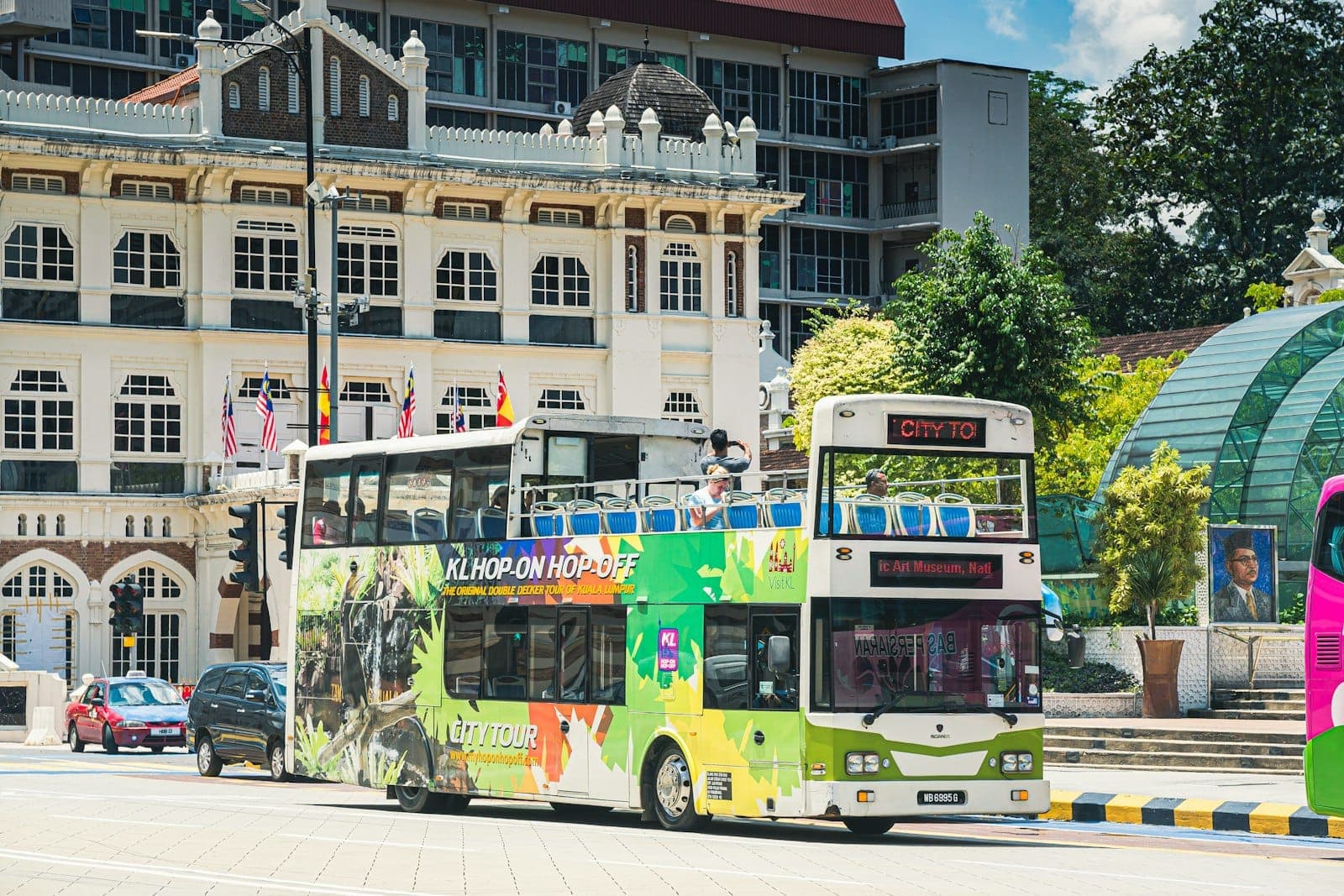 Bus tingkat hop-on hop-off Kuala Lumpur yang berwarna-warni melewati gedung-gedung kolonial dan bendera Malaysia di jalan kota yang cerah.