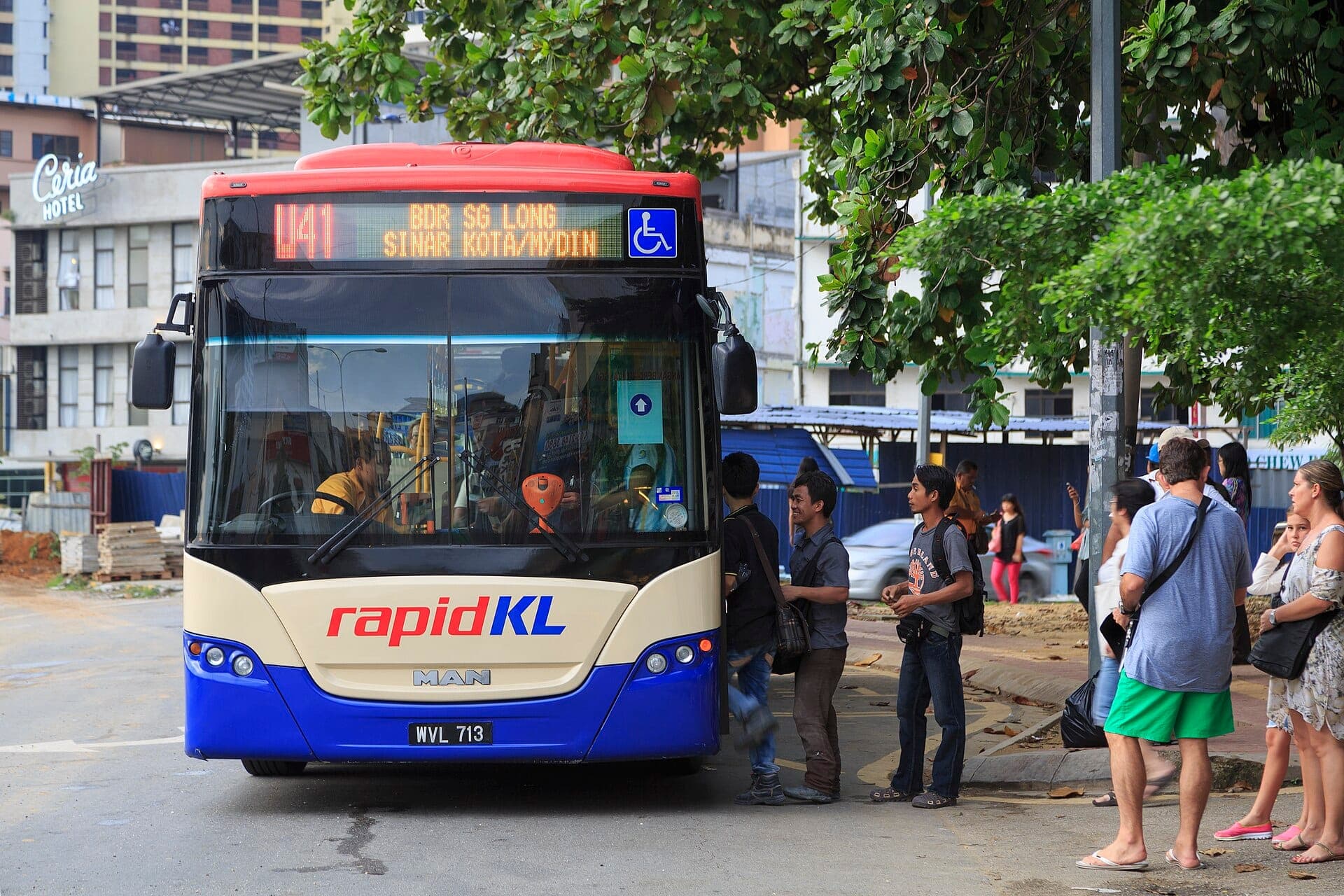 Orang-orang mengantri untuk naik bus kota RapidKL di bawah pohon-pohon rindang di pusat kota Kuala Lumpur, dengan latar belakang gedung-gedung dan proyek konstruksi perkotaan.