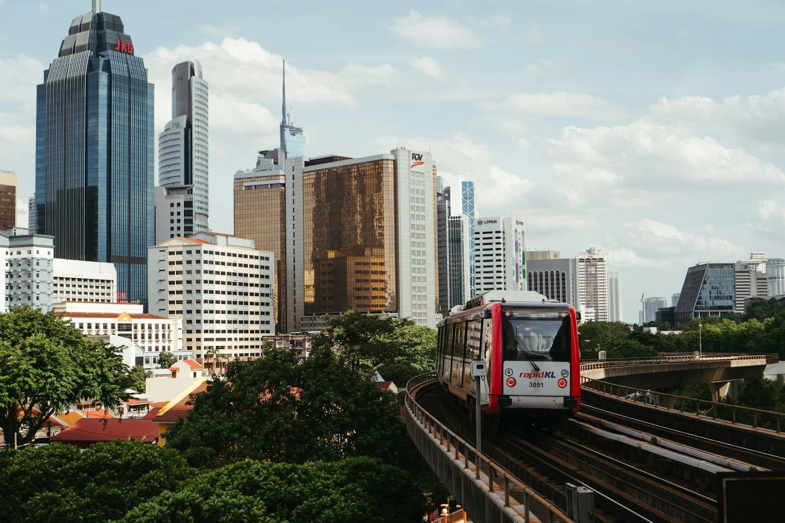 Kereta Rapid KL melaju di jalur layang dengan pemandangan cakrawala Kuala Lumpur, gedung pencakar langit modern, dan pepohonan hijau yang rimbun di bawah langit yang sebagian berawan.