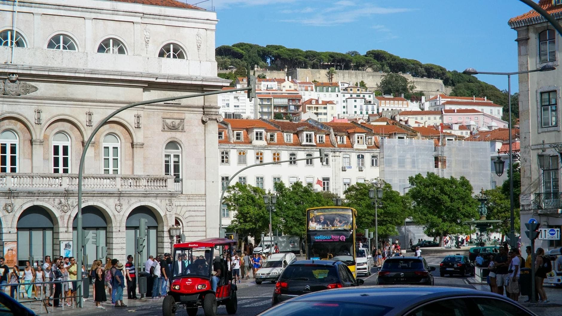 Rua movimentada de Lisboa com um autocarro urbano, pessoas esperando na paragem e edifícios clássicos ao fundo, sugerindo transporte urbano.