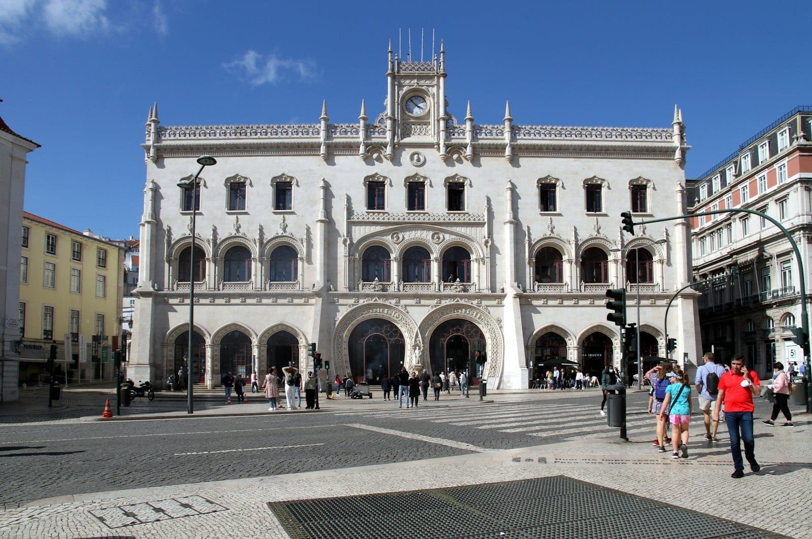 Vista frontal da estação de comboios do Rossio em Lisboa com pedestres atravessando na frente em um dia claro.