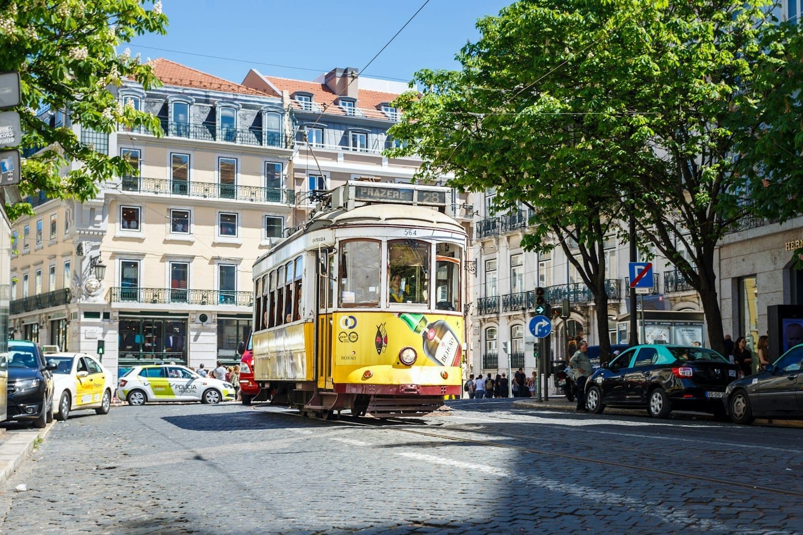 O icônico bonde amarelo passando por uma charmosa rua de Lisboa repleta de prédios históricos, árvores e táxis sob a luz intensa do dia, capturando o clima de transporte da cidade.