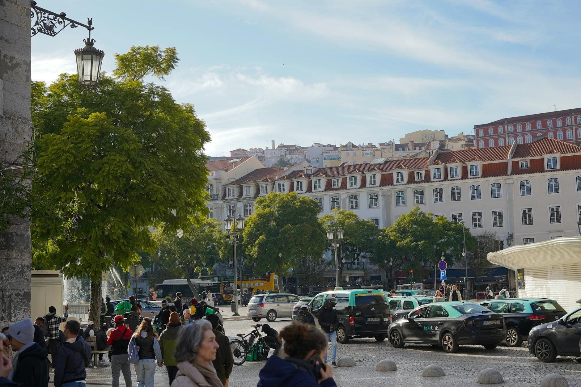 Rua de Lisboa com vários táxis e carros estacionados ao longo do passeio, pessoas caminhando e edifícios portugueses ao fundo.