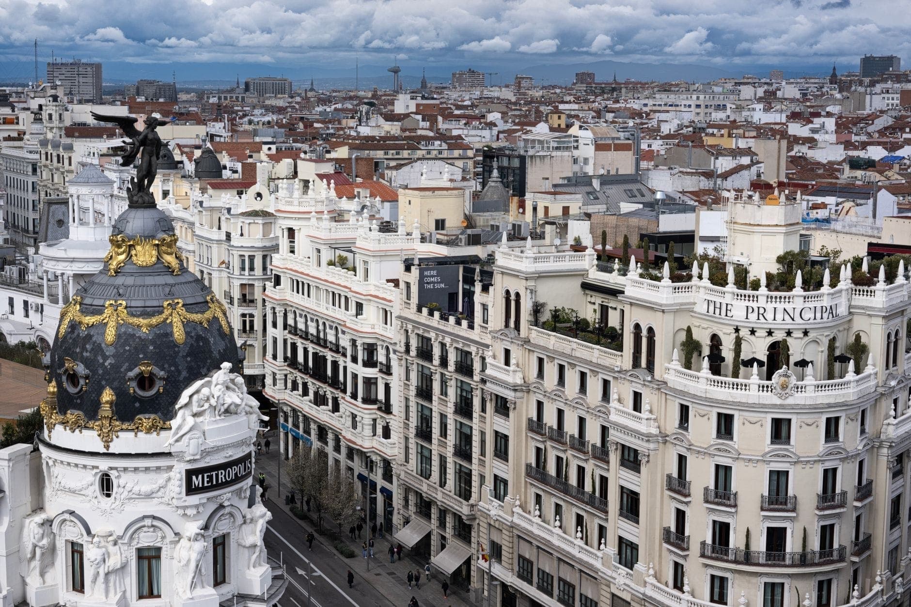Vista aérea de la Gran Vía de Madrid con el icónico edificio Metropolis, calles concurridas y paisaje urbano bajo cielos nublados.