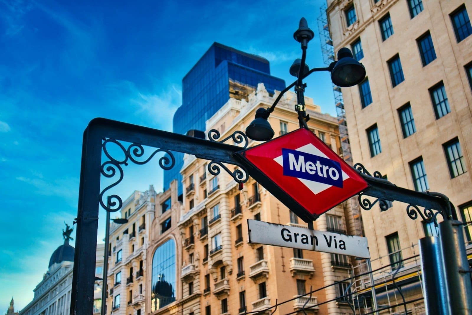 Señal del Metro de Madrid en la estación de Gran Vía con edificios históricos al fondo en un día soleado.