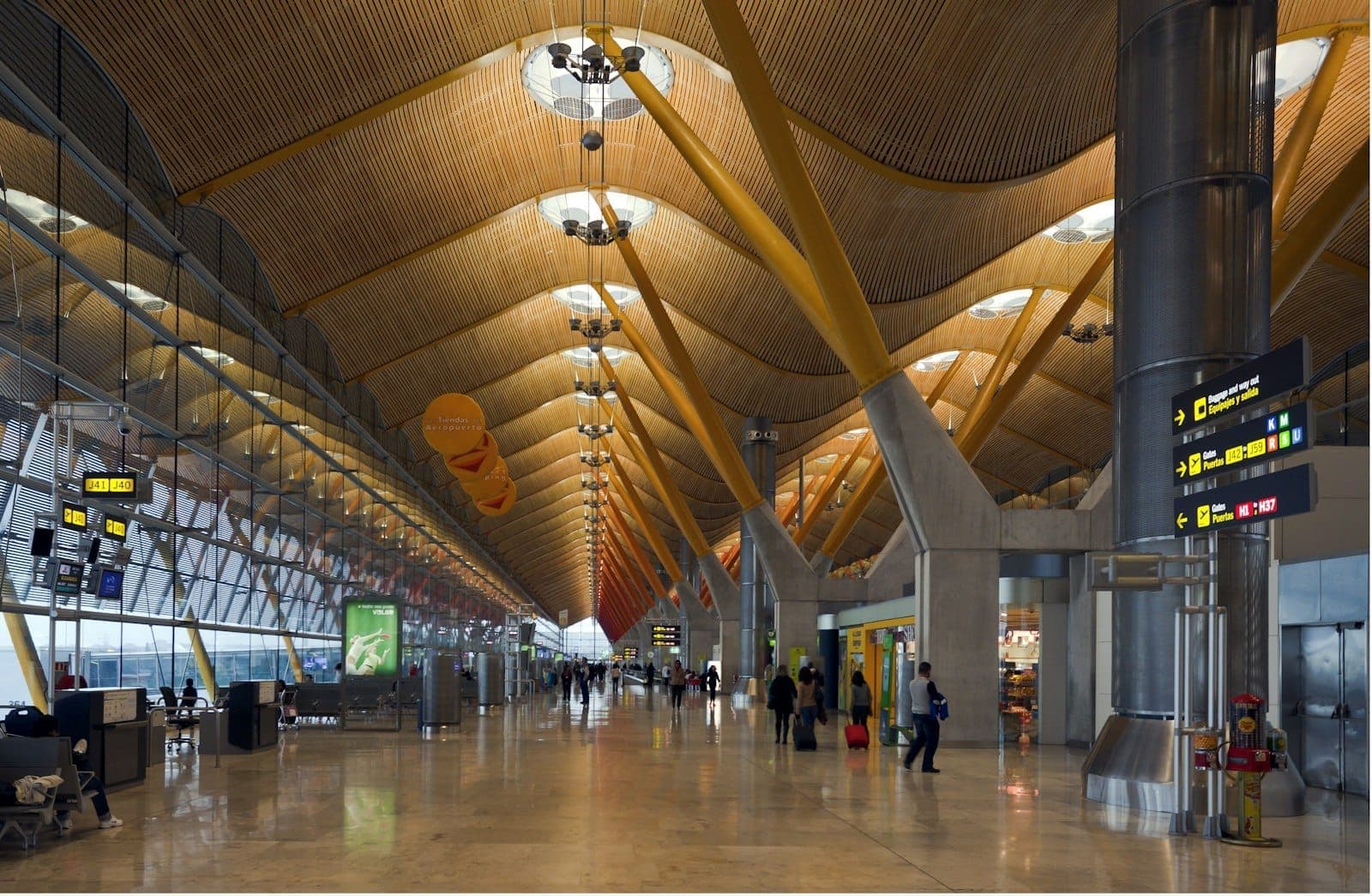 Interior espacioso de la terminal del Aeropuerto Madrid Barajas con vigas amarillas, señalización y viajeros caminando por el vestíbulo.