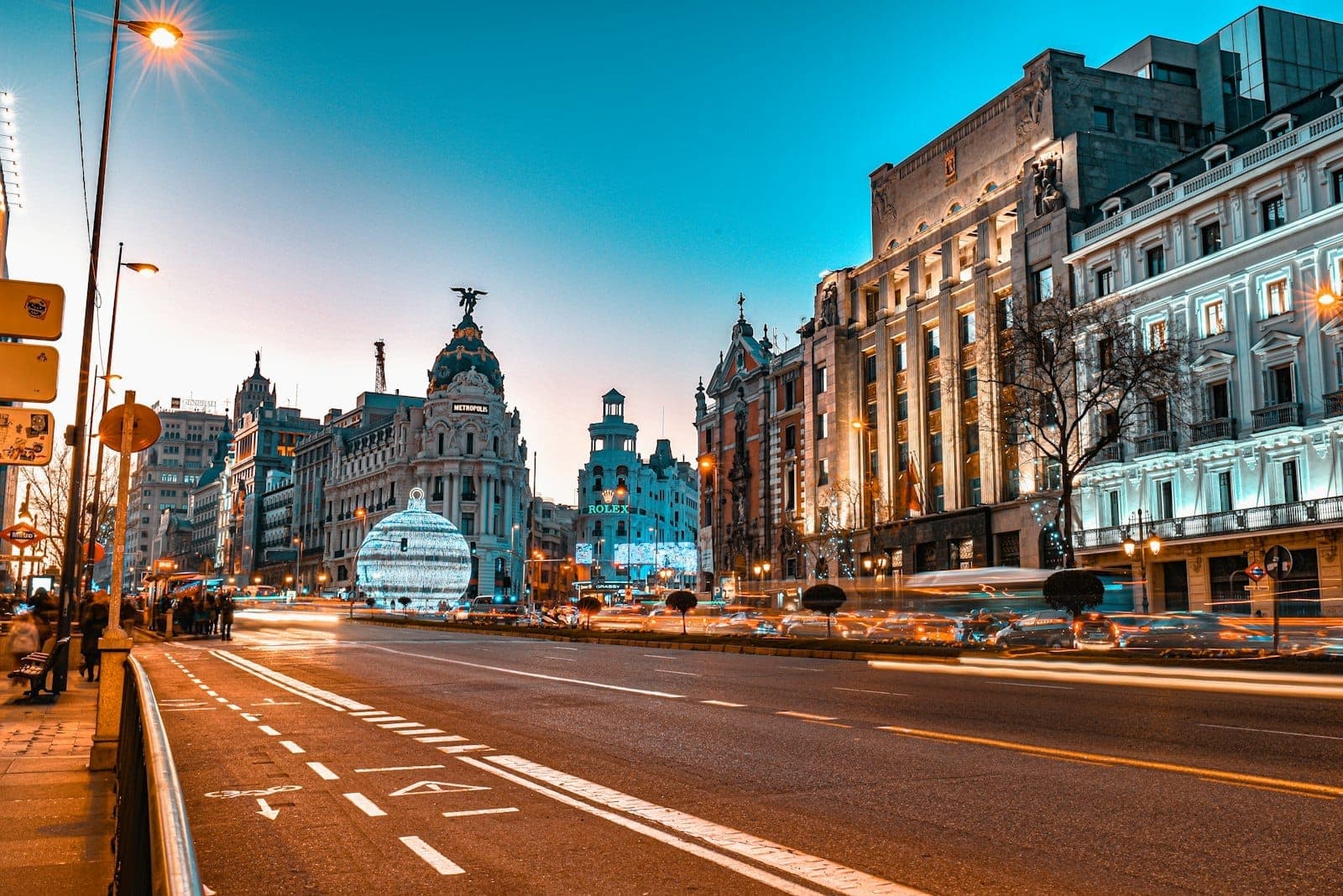 วิวถนน Gran Via ในมาดริดที่มีรถผ่านไปมาคึกคัก ท่ามกลางอาคารสไตล์ประวัติศาสตร์และท้องฟ้าแสงสนธยา สัมผัสบรรยากาศการเดินทางในเมืองที่มีชีวิตชีวา