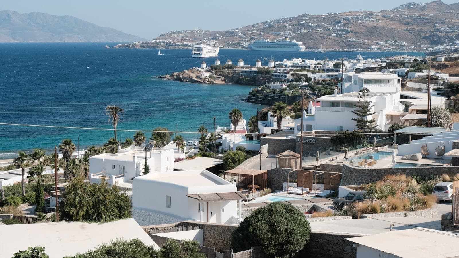 Panoramablick auf die Küste von Mykonos mit weißen Gebäuden, Kreuzfahrtschiff und Fähren, die im Hafen ankommen.