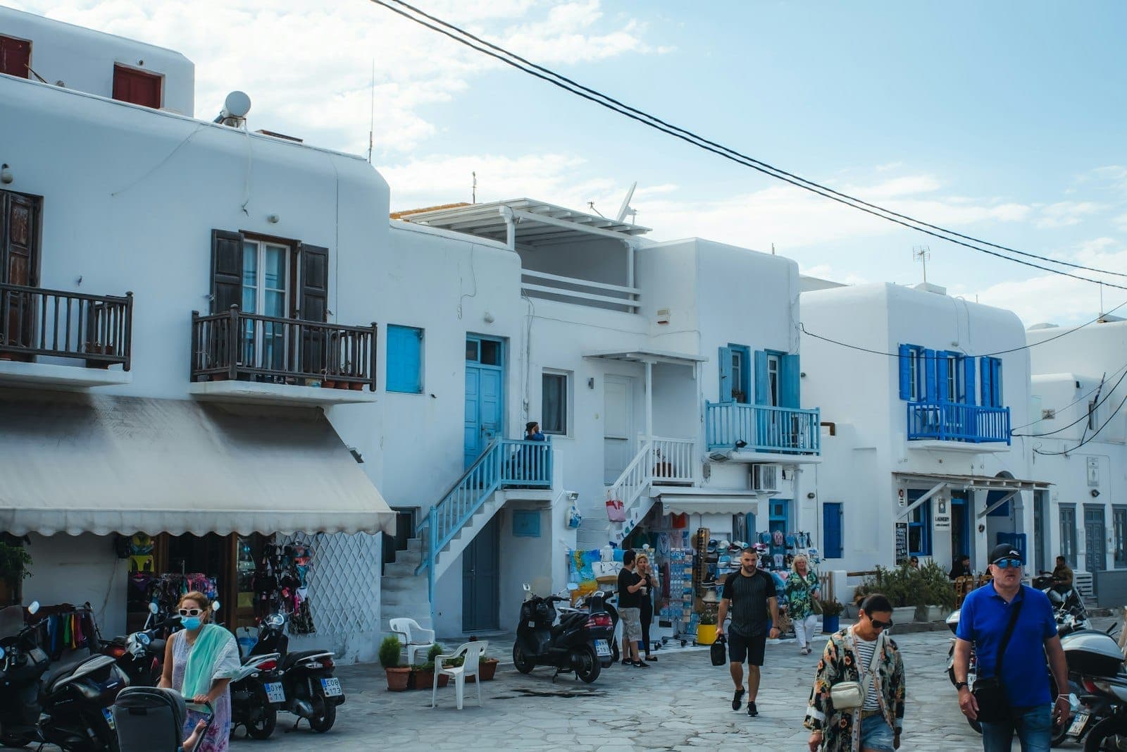 Menschen, die durch eine von weißen Gebäuden gesäumte Straße in Mykonos spazieren, mit geparkten Rollern und Motorrädern im Vordergrund.