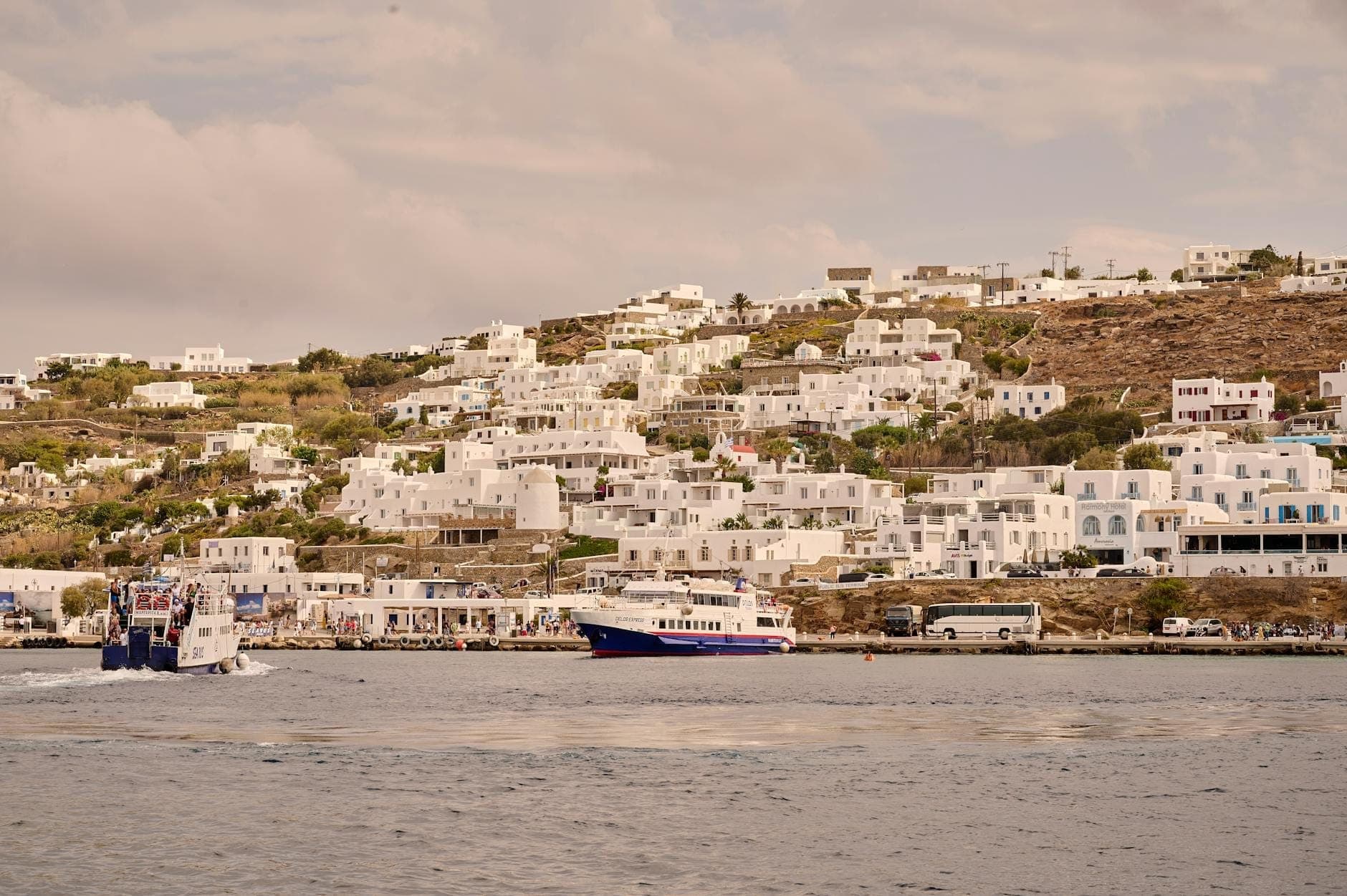 Zwei Passagierboote fahren zwischen dem Alten Hafen und den weiß gekalkten Gebäuden von Mykonos, mit Häusern am Hang im Hintergrund.