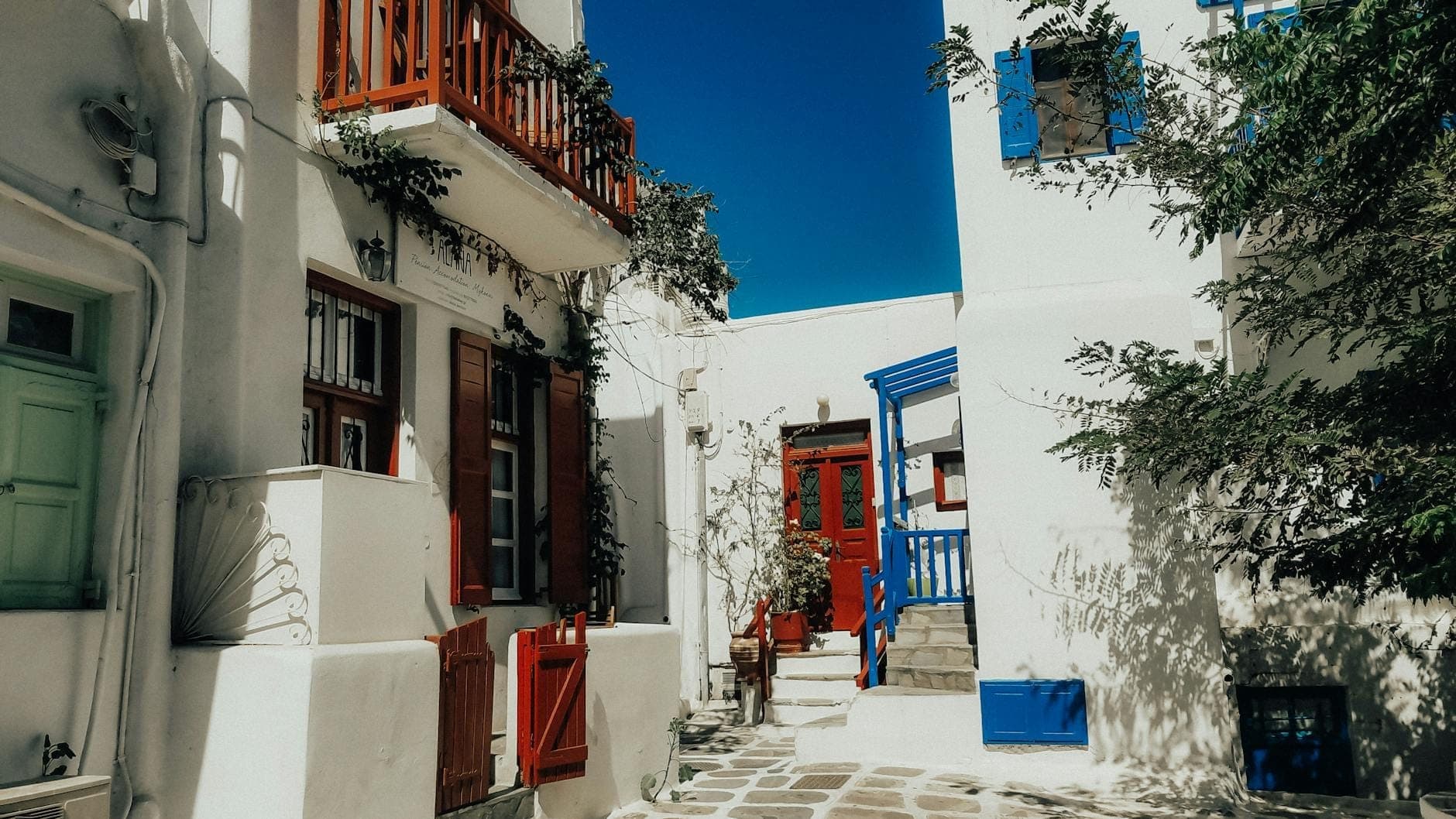 Typische Gasse in Mykonos Town mit weiß getünchten Gebäuden, bunten Türen und Fensterläden, schmalem Steinweg und blauem Himmel.