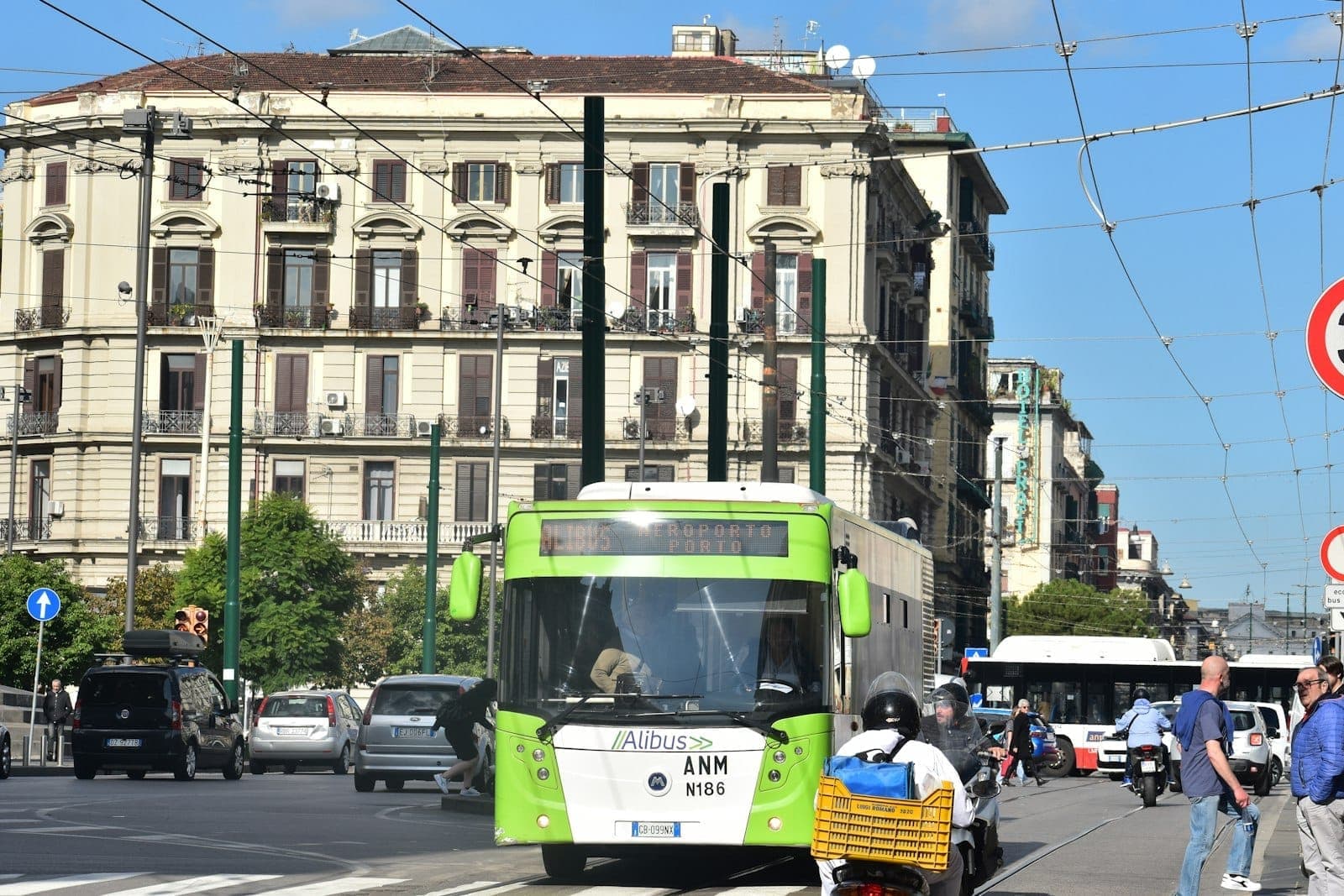 Ônibus verde do transporte público Alibus no centro de Nápoles, com pessoas, carros e prédios históricos ao fundo em um dia ensolarado.