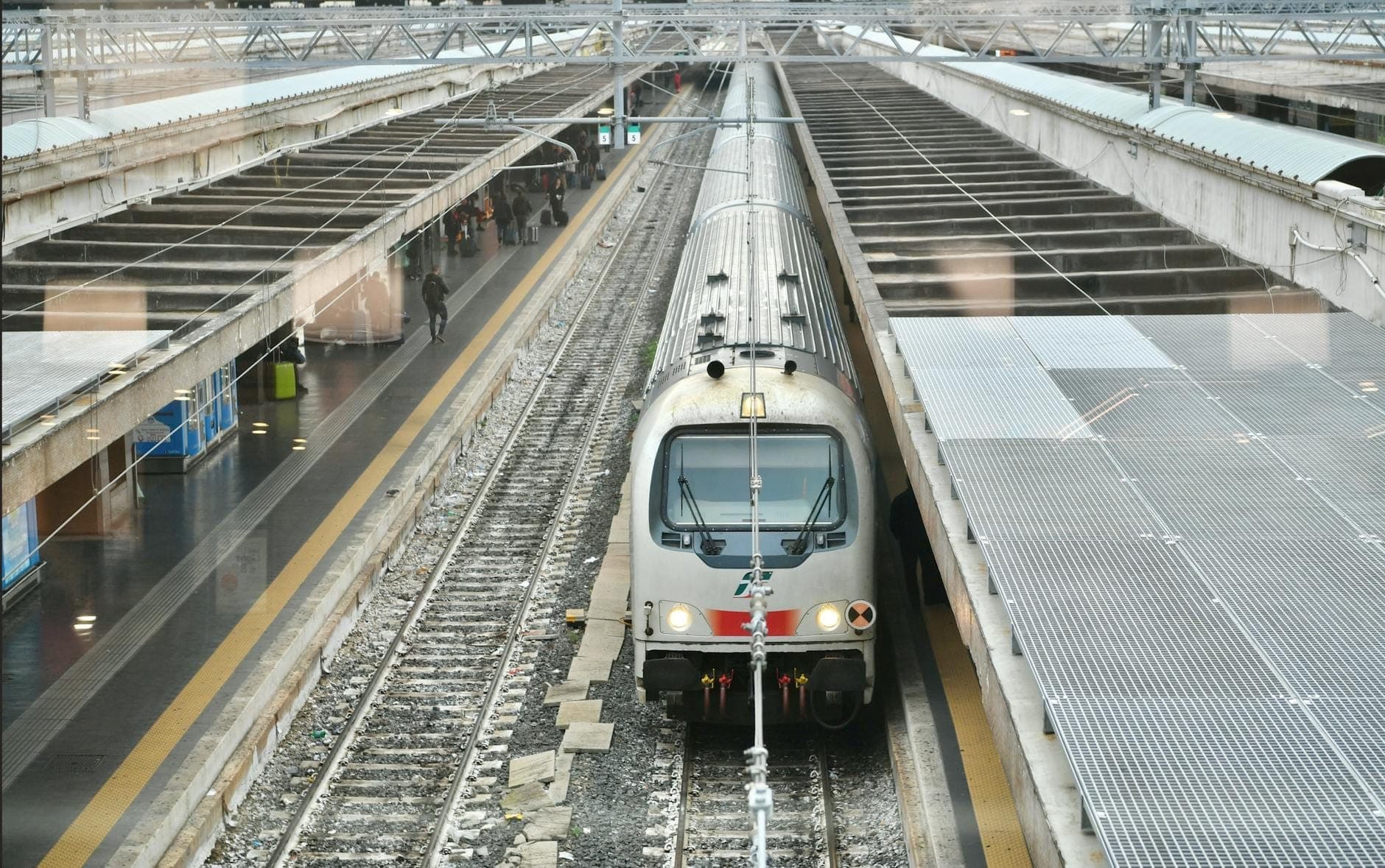 Um moderno trem regional prateado chega a uma grande estação de trem aberta em Nápoles, com plataformas cobertas e viajantes esperando nas proximidades.