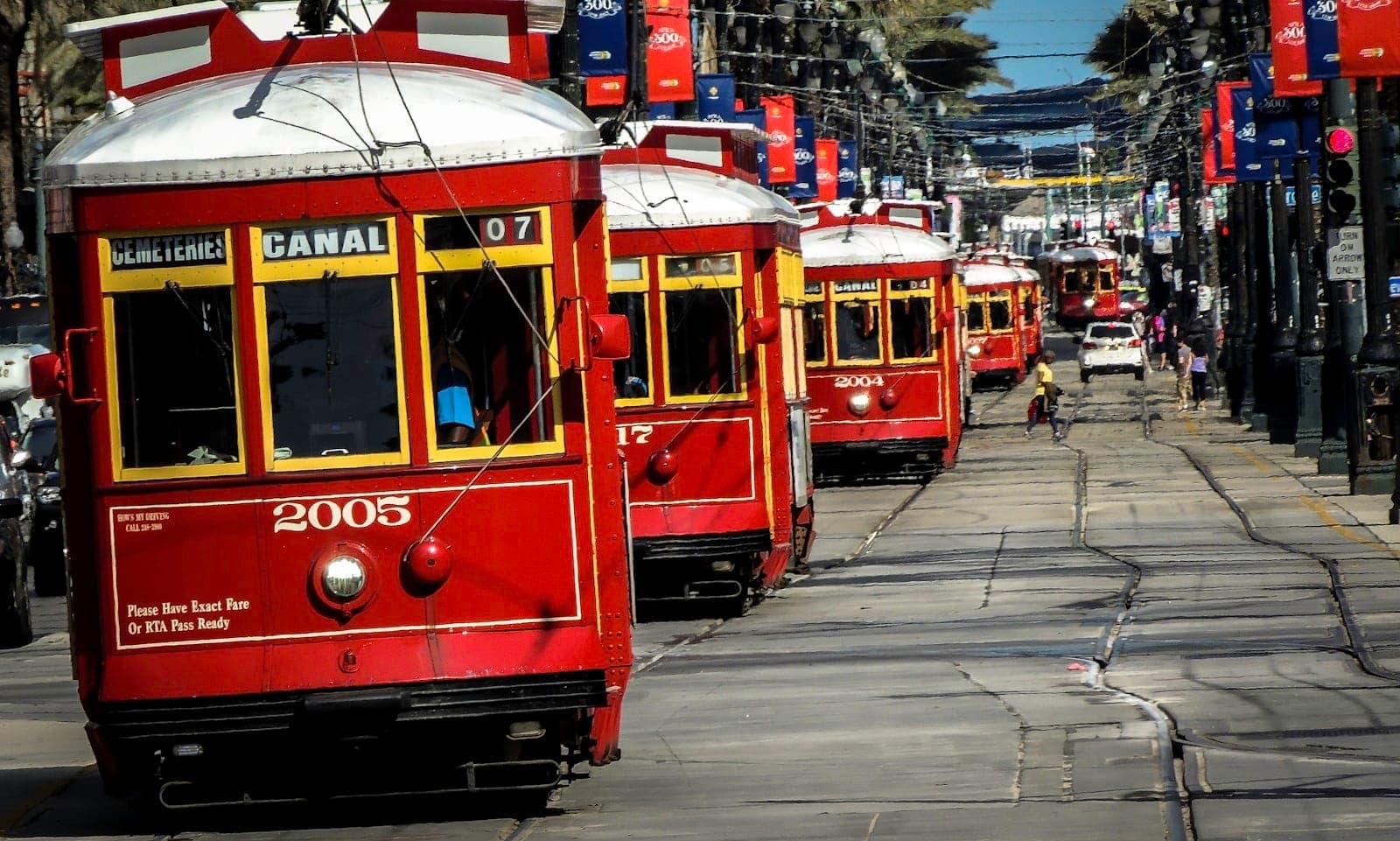 รถรางสีแดงคลาสสิกหลายคันของ New Orleans เรียงรายอยู่บน Canal Street มีต้นปาล์มและป้ายแบนเนอร์ให้เห็น สร้างบรรยากาศการเดินทางในเมืองที่คึกคัก