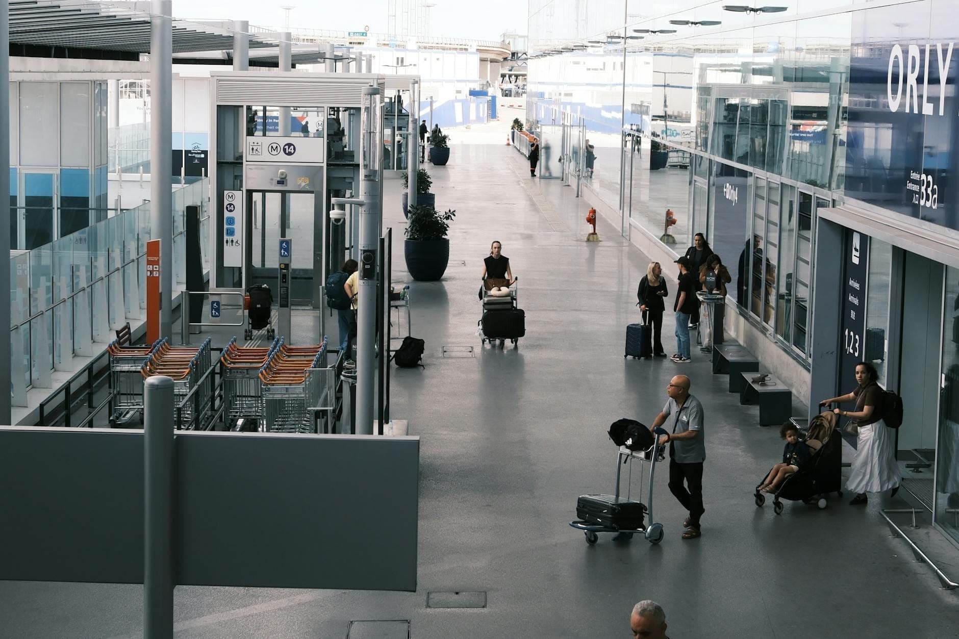 Exterior view of Paris Orly airport with people, luggage carts, and glass terminal entrance visible, clearly marked with 'ORY' signage.