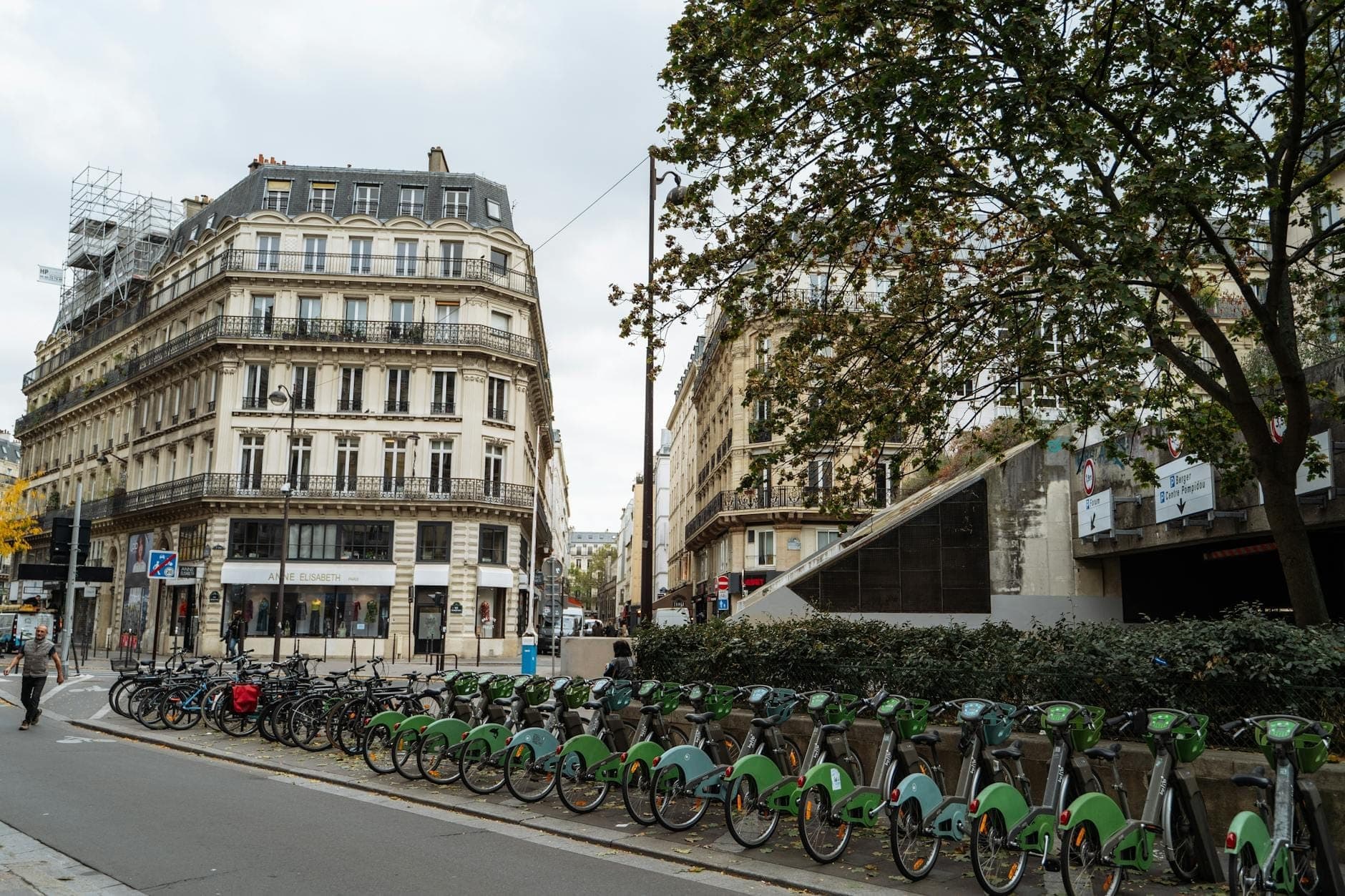 A long row of green Vélib' bikes lined up at a docking station on a Paris street, with classic Parisian buildings and trees visible in the background.