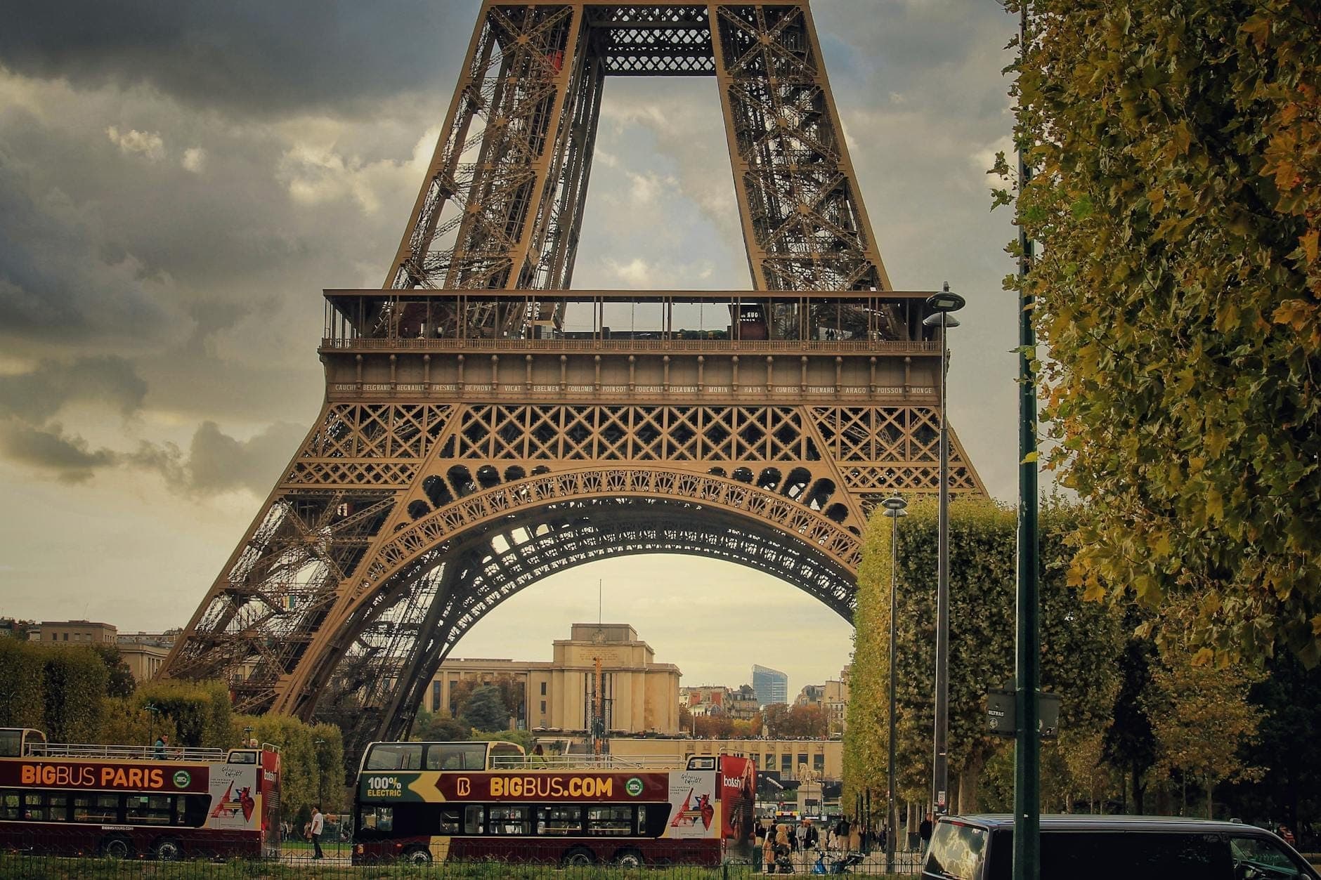 Two Paris city tour buses driving beneath the Eiffel Tower on a city street, with trees lining the road.