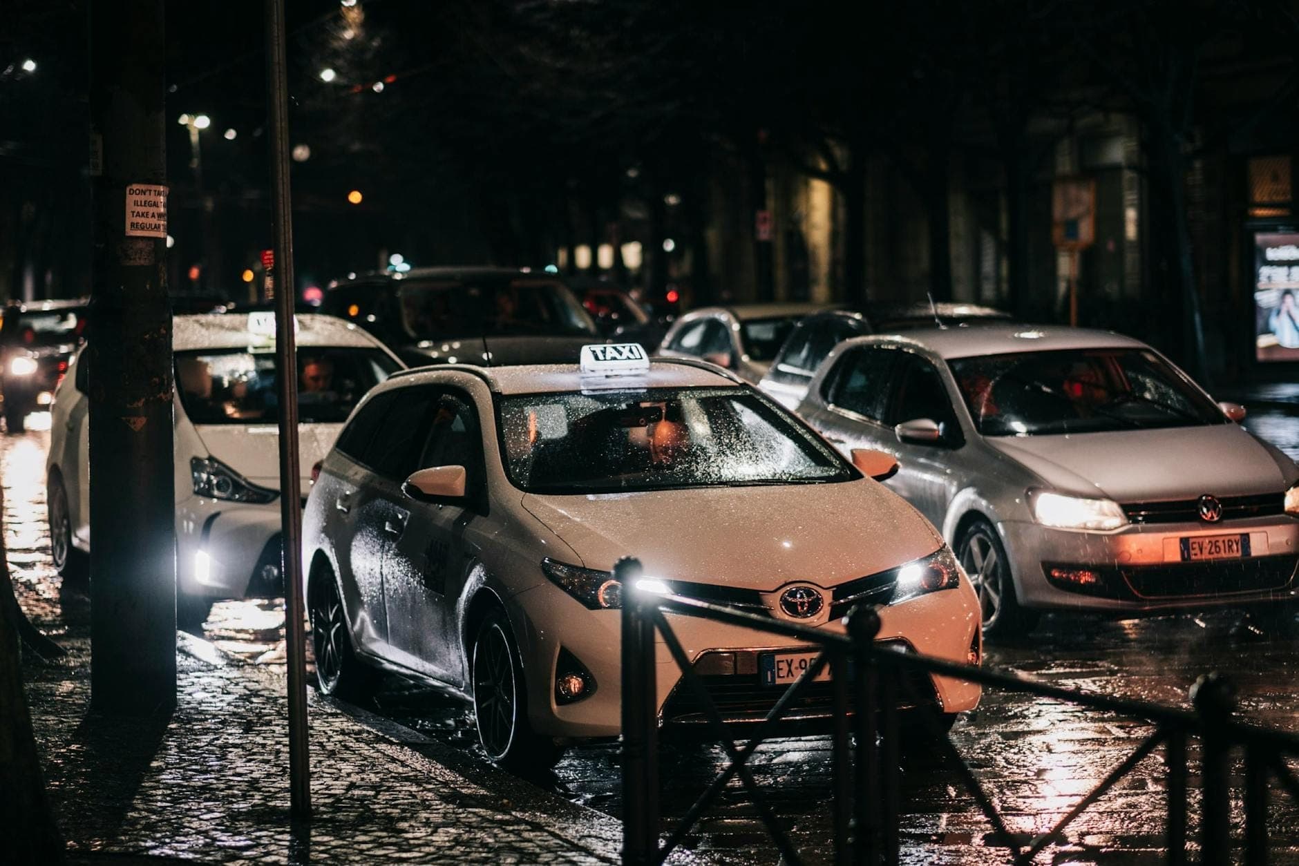White taxi with illuminated roof sign waits in traffic on a rainy Parisian street at night, surrounded by other cars.