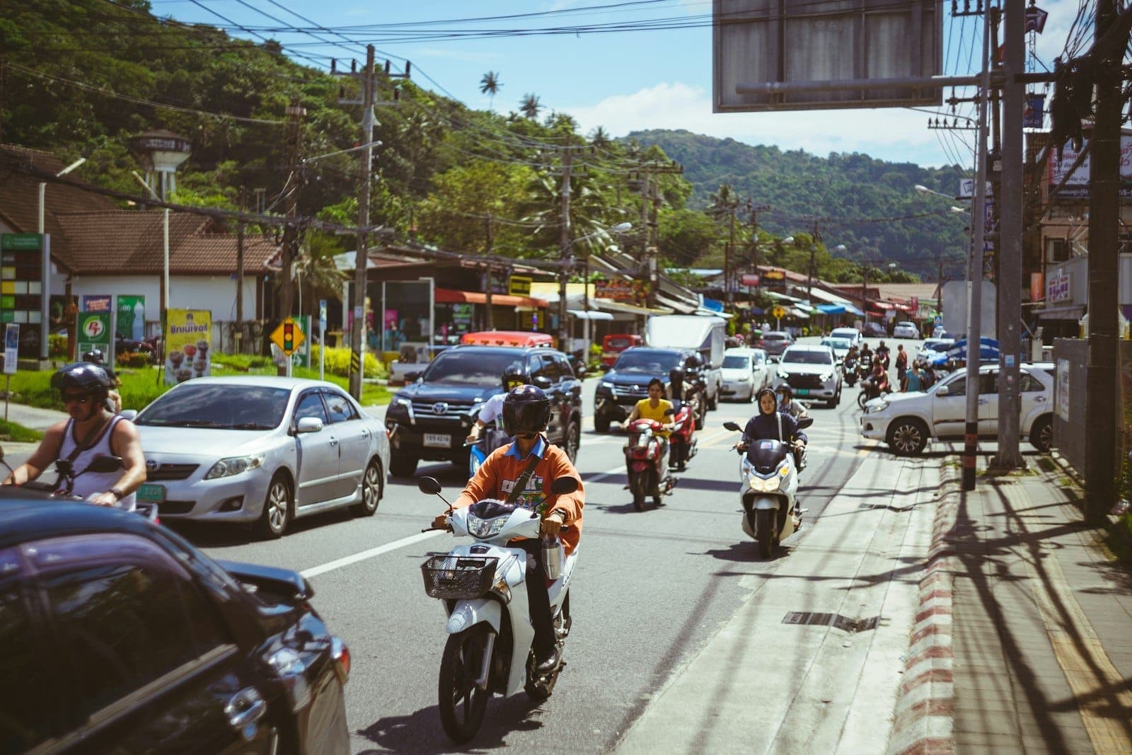 Busy Phuket street with cars and motorbikes in traffic, shops and green hills in background, showing typical island transport options.