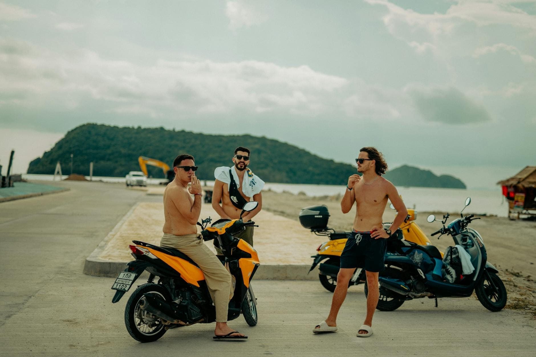 Tres personas con scooters cerca de una carretera costera con colinas y océano al fondo, típico del estilo de transporte popular en Phuket.