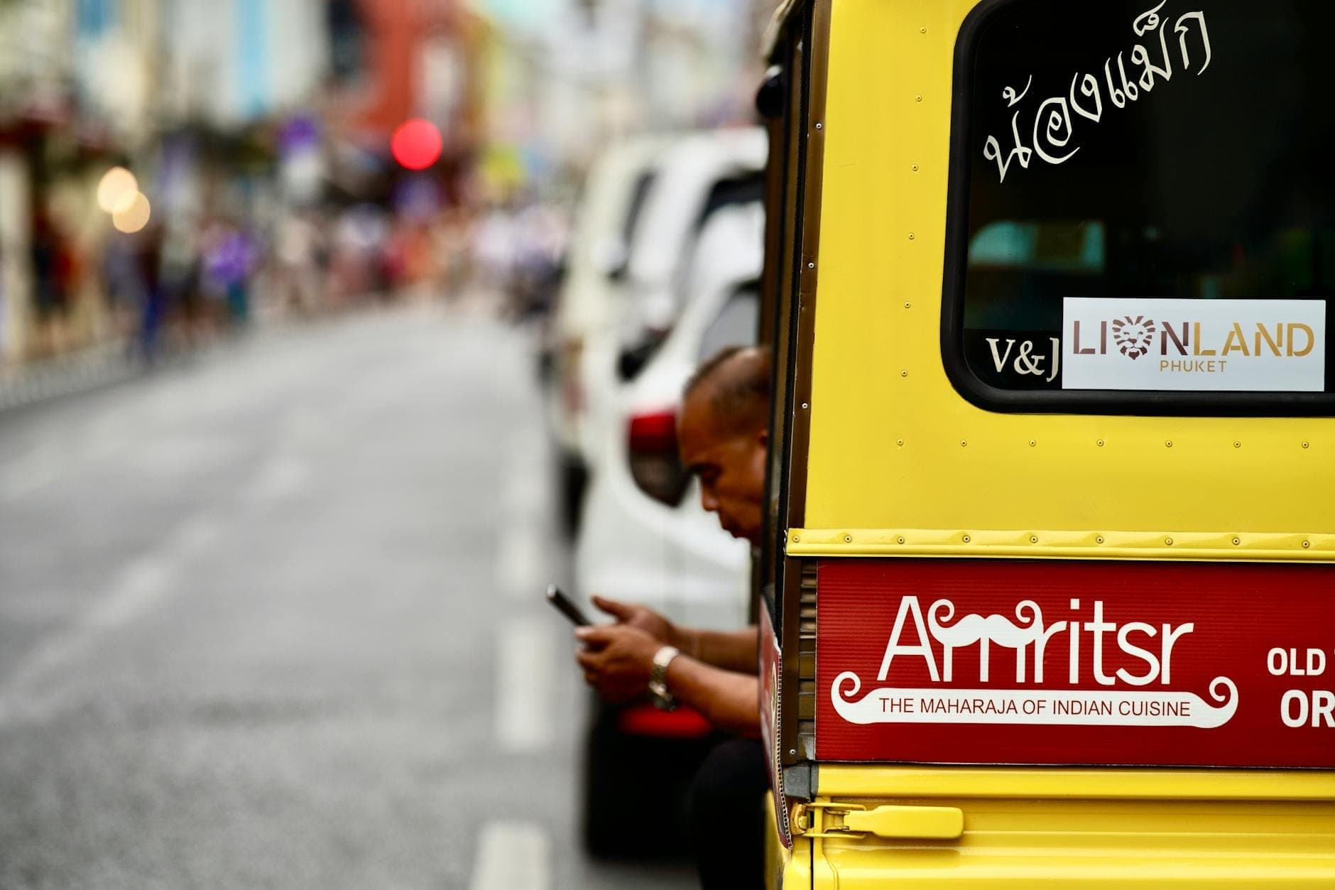 Un tuk-tuk estilo camioneta amarillo con un hombre dentro, estacionado en una calle del casco antiguo de Phuket.
