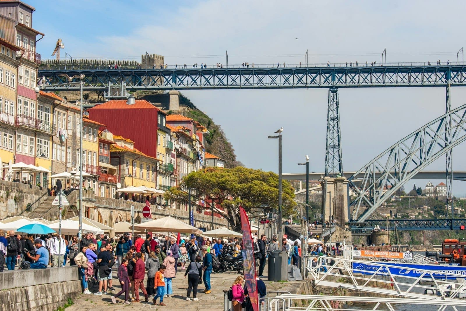 Folle di persone camminano lungo l'animato lungofiume di Porto con edifici colorati e il Ponte Dom Luís I sullo sfondo in una giornata di sole.