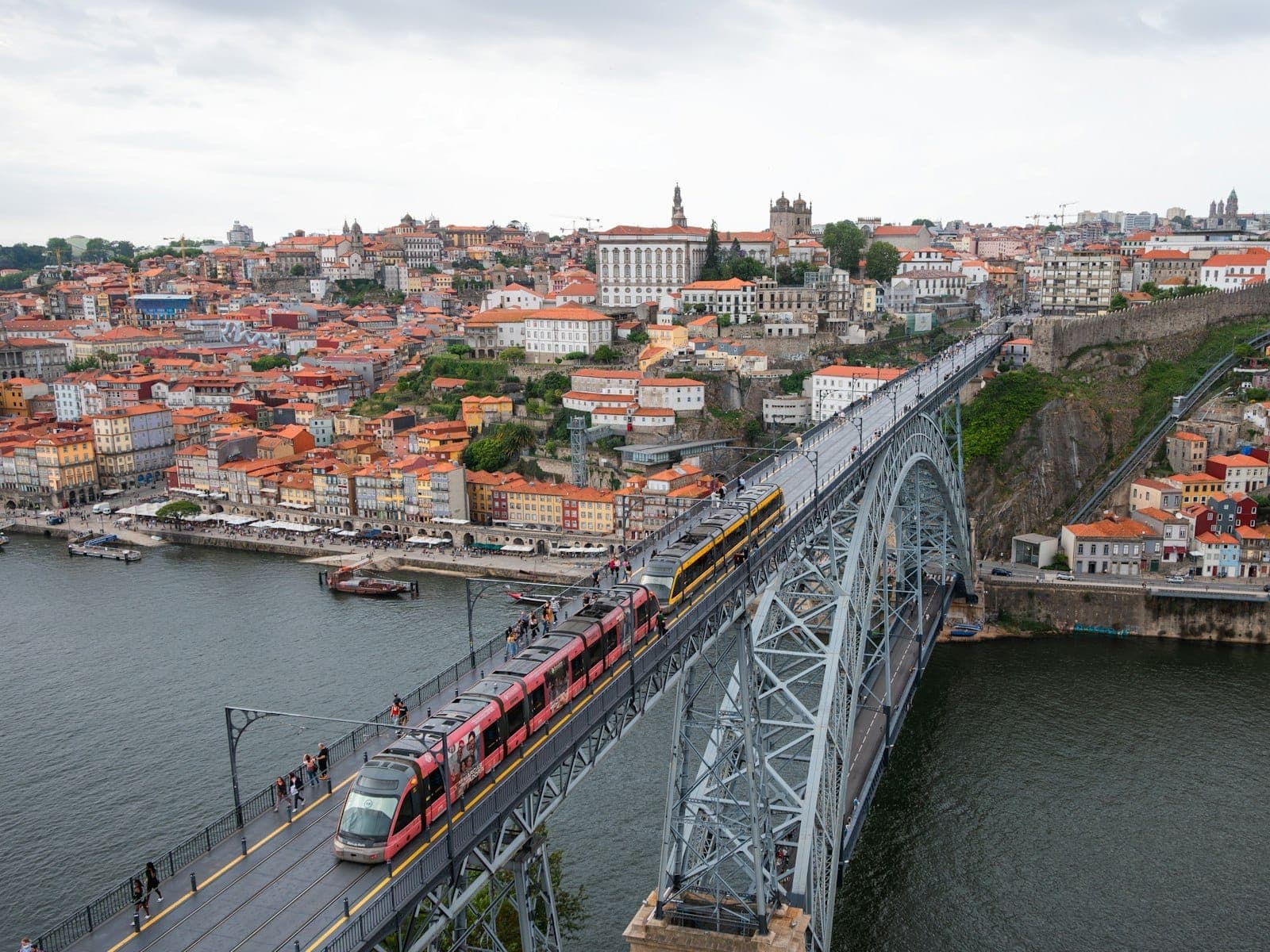 Un tram moderno e un classico tram giallo attraversano il Ponte Dom Luís I sopra il lungofiume storico di Porto con lo skyline della città sullo sfondo.
