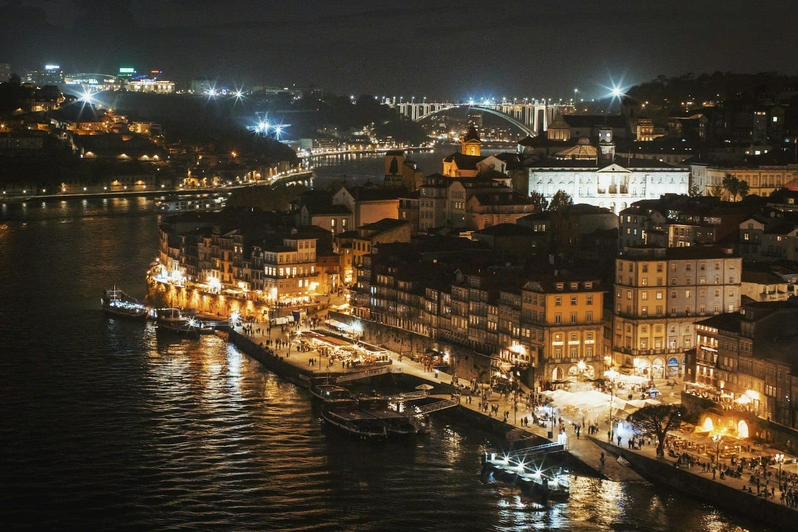 Vista notturna dello storico lungofiume di Porto con edifici, luci vivaci e la città riflessa sul fiume Douro.