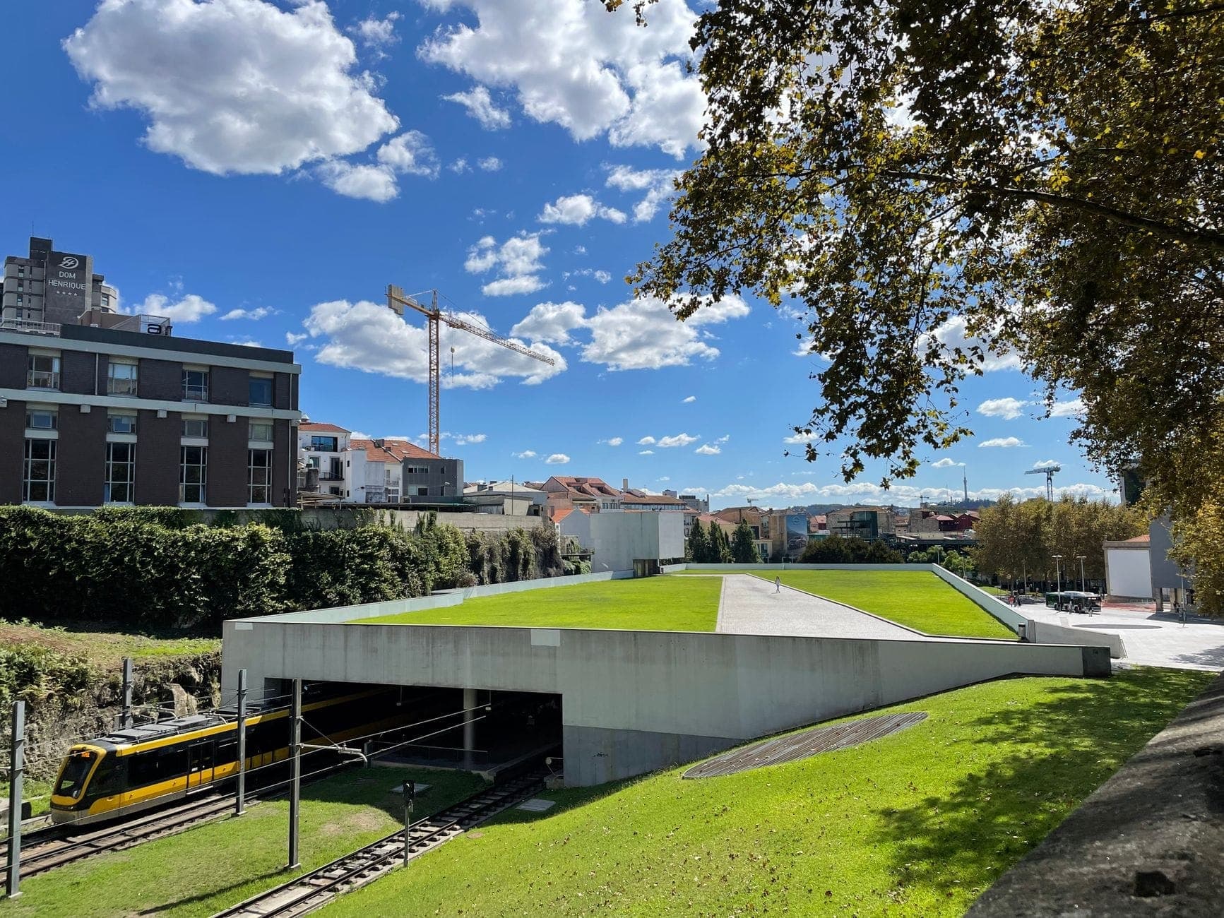 Vista esterna del treno della metropolitana leggera Porto Metro che emerge dall'ingresso di una galleria moderna sotto un tetto erboso, con gli edifici della città sullo sfondo.