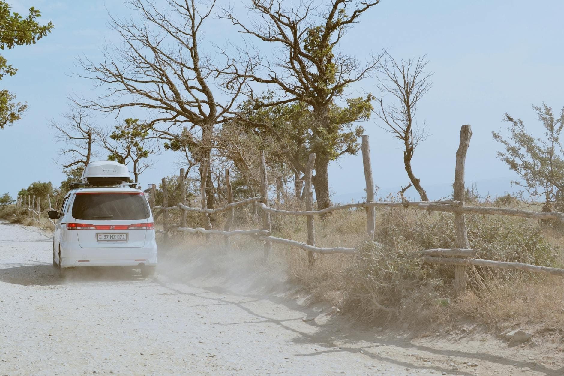 Car driving on a dusty rural road lined with rustic wooden fences and sparse trees under a bright sky.