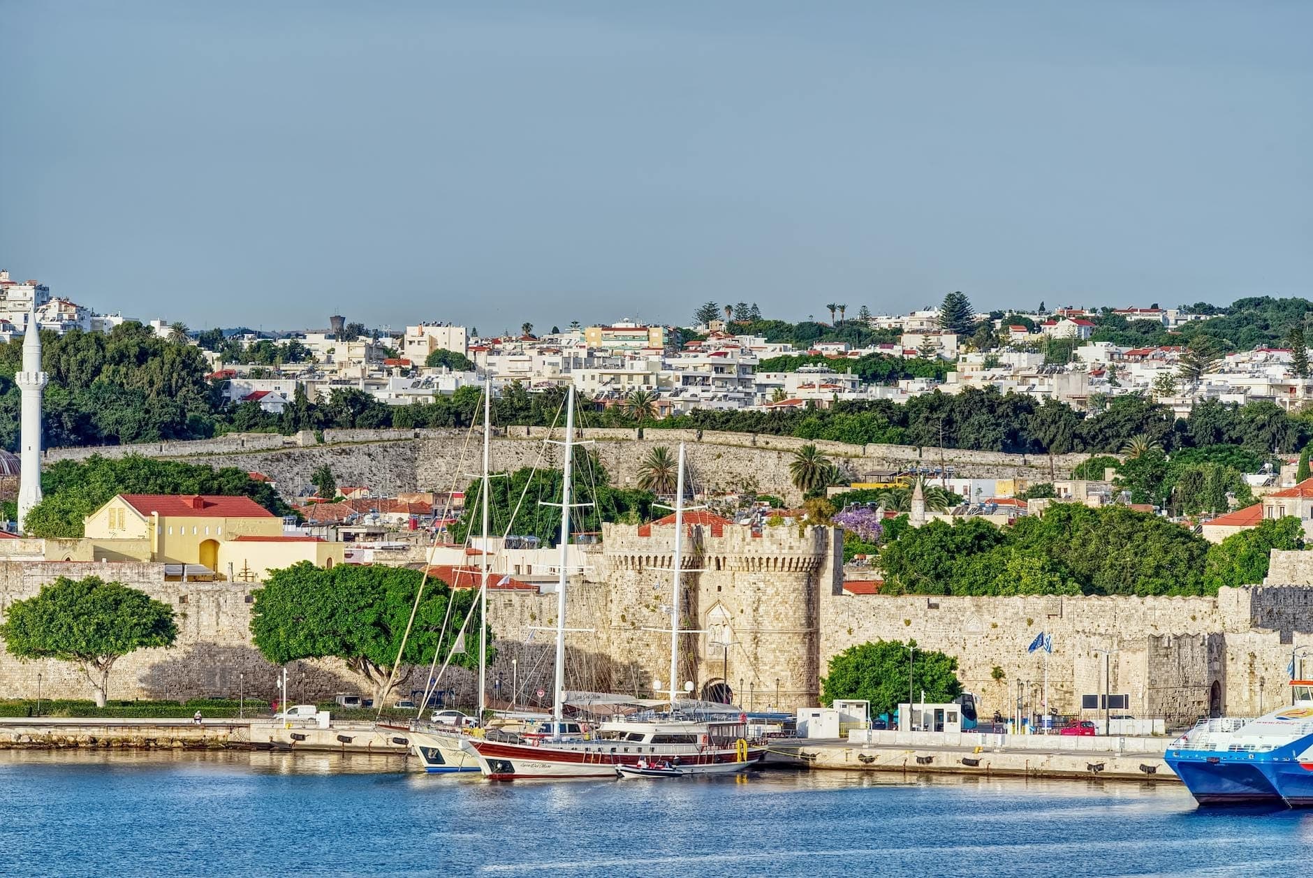 Vue sur la ville de Rhodes avec ses remparts en pierre, des voiliers amarrés dans un port aux eaux bleu vif, et des maisons à flanc de colline sous un ciel dégagé.