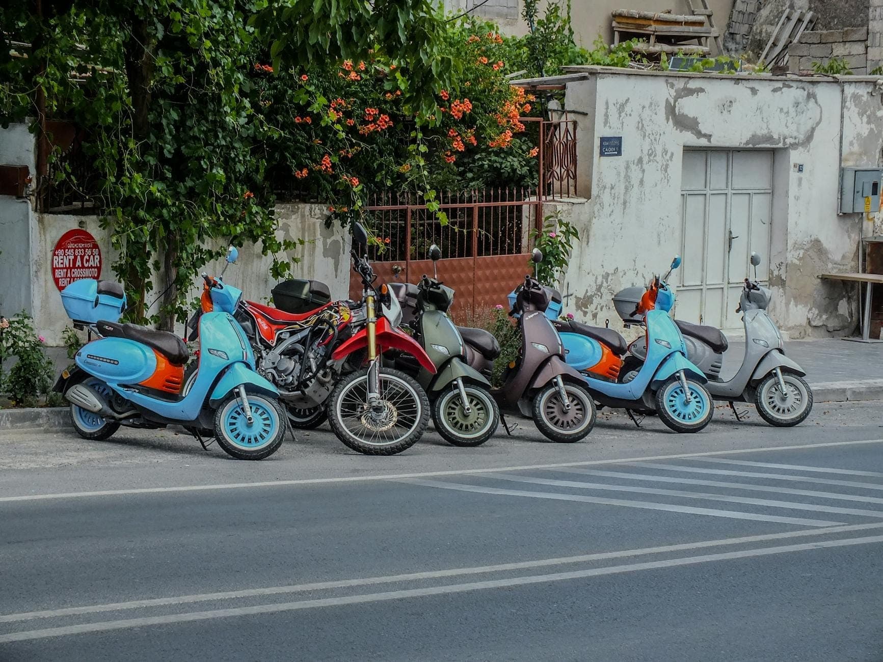 Several colorful scooters and a motorcycle parked by the roadside in a Greek village with whitewashed buildings and flowers.