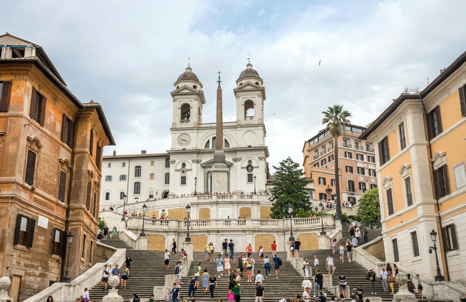 Góc nhìn rộng của Spanish Steps ở Rome với người đi bộ qua lại, được bao quanh bởi các tòa nhà lịch sử dưới bầu trời nhiều mây.