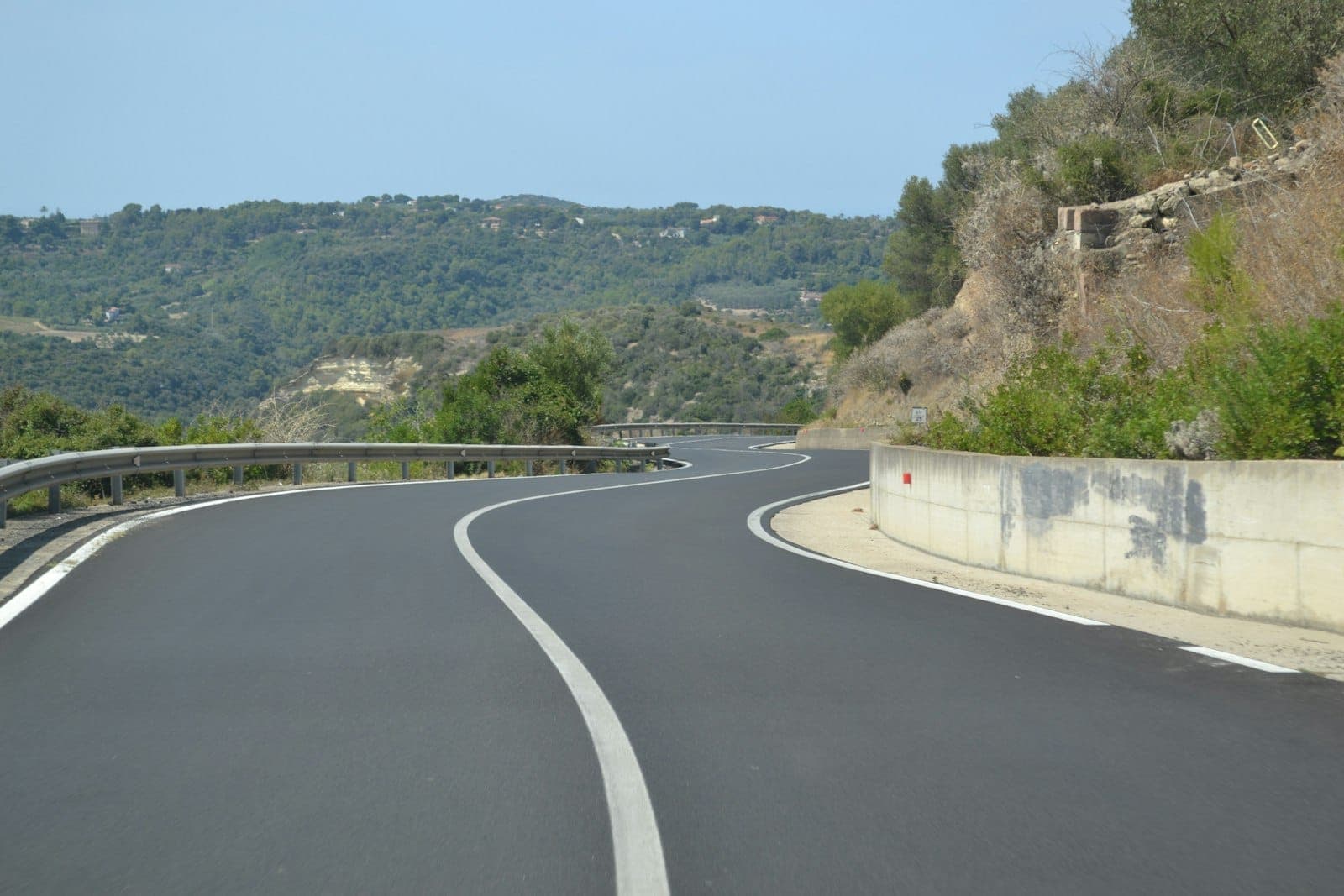 Tramo curvo de una carretera sarda bien conservada con colinas y vegetación al fondo bajo un cielo despejado.