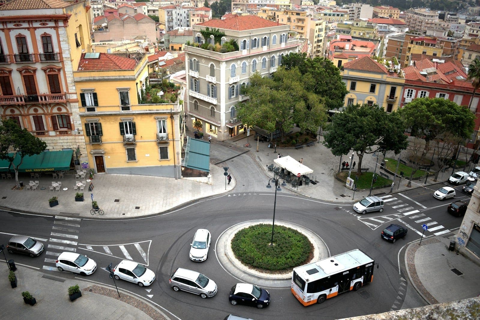 Una rotonda en el centro de una ciudad de Cerdeña con taxis, coches y un autobús circulando por las calles, rodeada de coloridos edificios históricos.