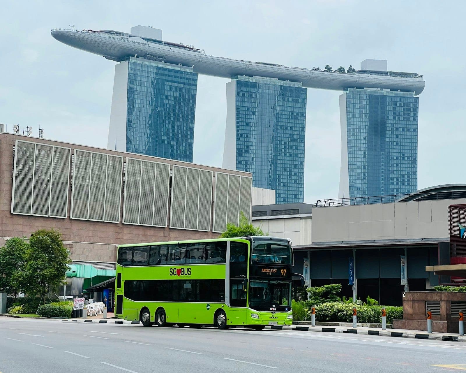 Ein leuchtend grüner Doppeldeckerbus aus Singapur vor dem Hotel Marina Bay Sands, klarer Himmel und Stadtgebäude im Hintergrund.