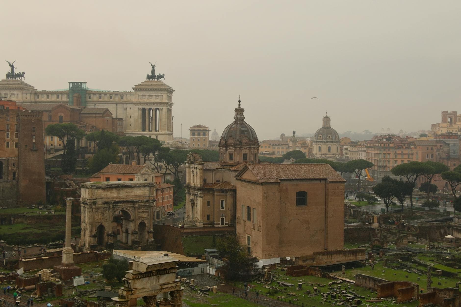 Panoramikong tanawin mula sa Gianicolo Hill na nagtatampok ng skyline ng Roma, mga sinaunang guho, mga kupolang simbahan, at ang monumento ng Vittoriano sa ilalim ng maulap na langit.