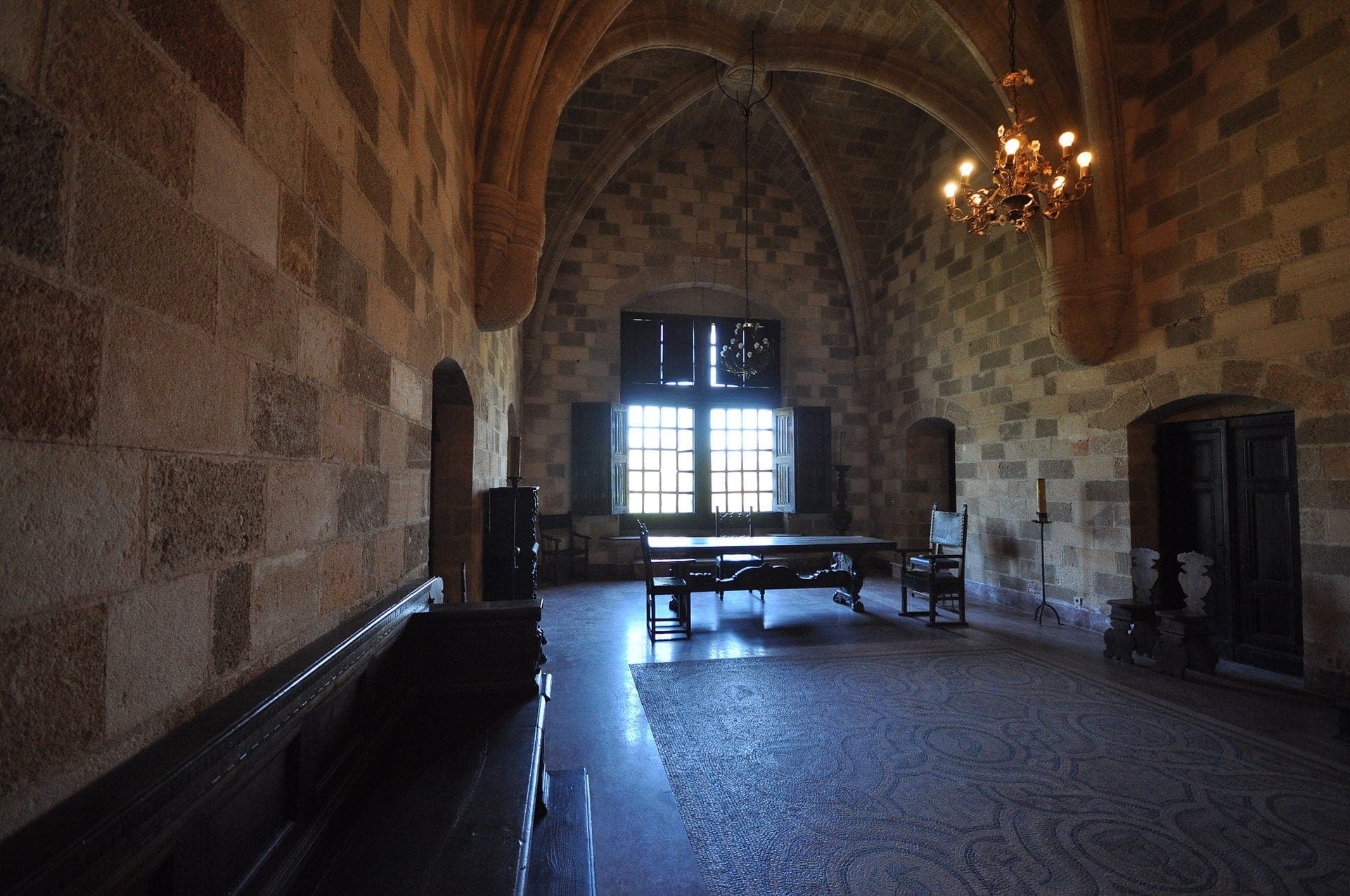Salle de pierre voûtée dans le Palais du Grand Maître de Rhodes, avec une architecture médiévale, des plafonds en arc, un sol en mosaïque, du mobilier en bois et un éclairage naturel doux.